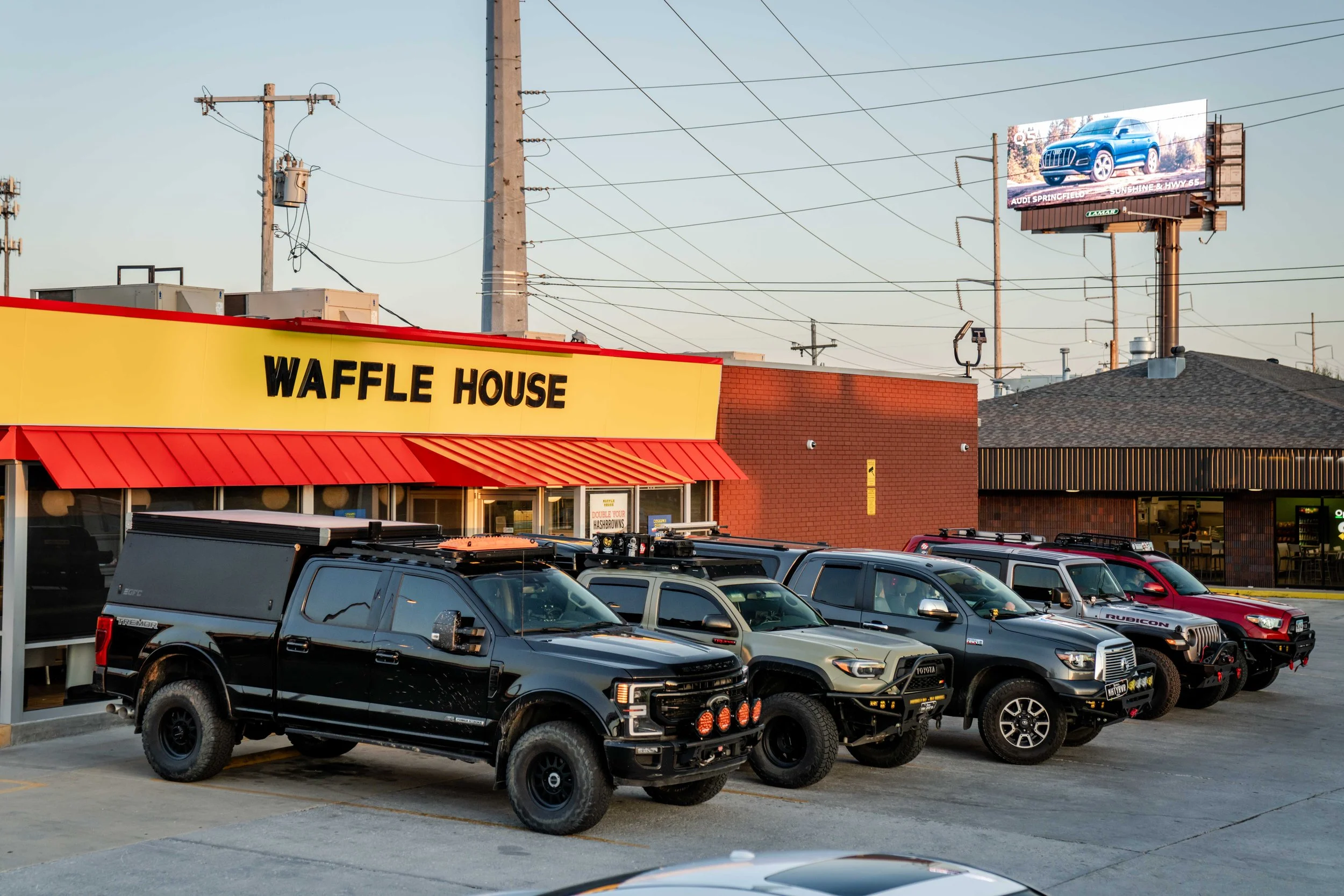 Parking lot with several black and gray pickup trucks parked in front of a Waffle House restaurant. A large billboard advertising an Audi SUV, and utility poles with power lines are visible in the background.