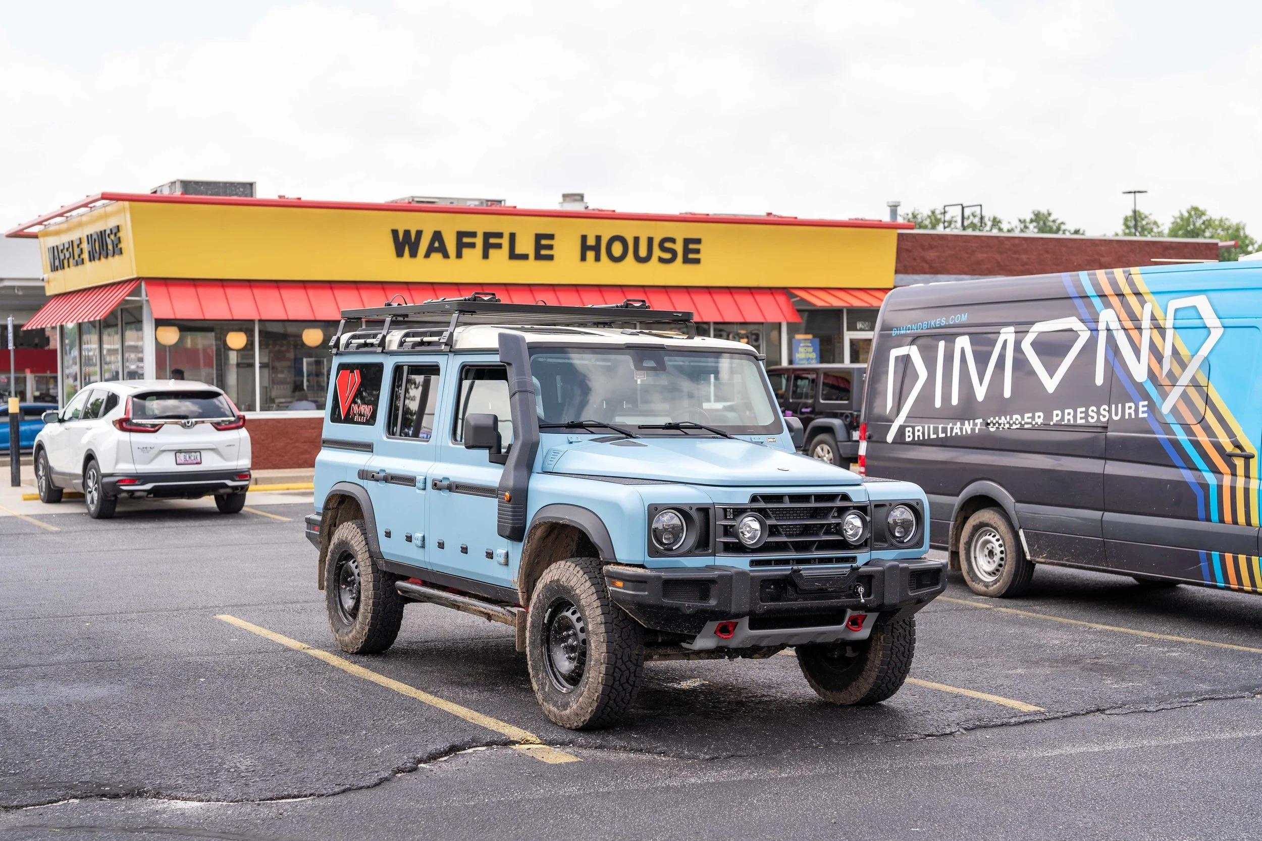 Parking lot scene with a Waffle House restaurant in the background, several cars parked, and a blue Ineos Grenadier and a branded van in the foreground, overcast sky.