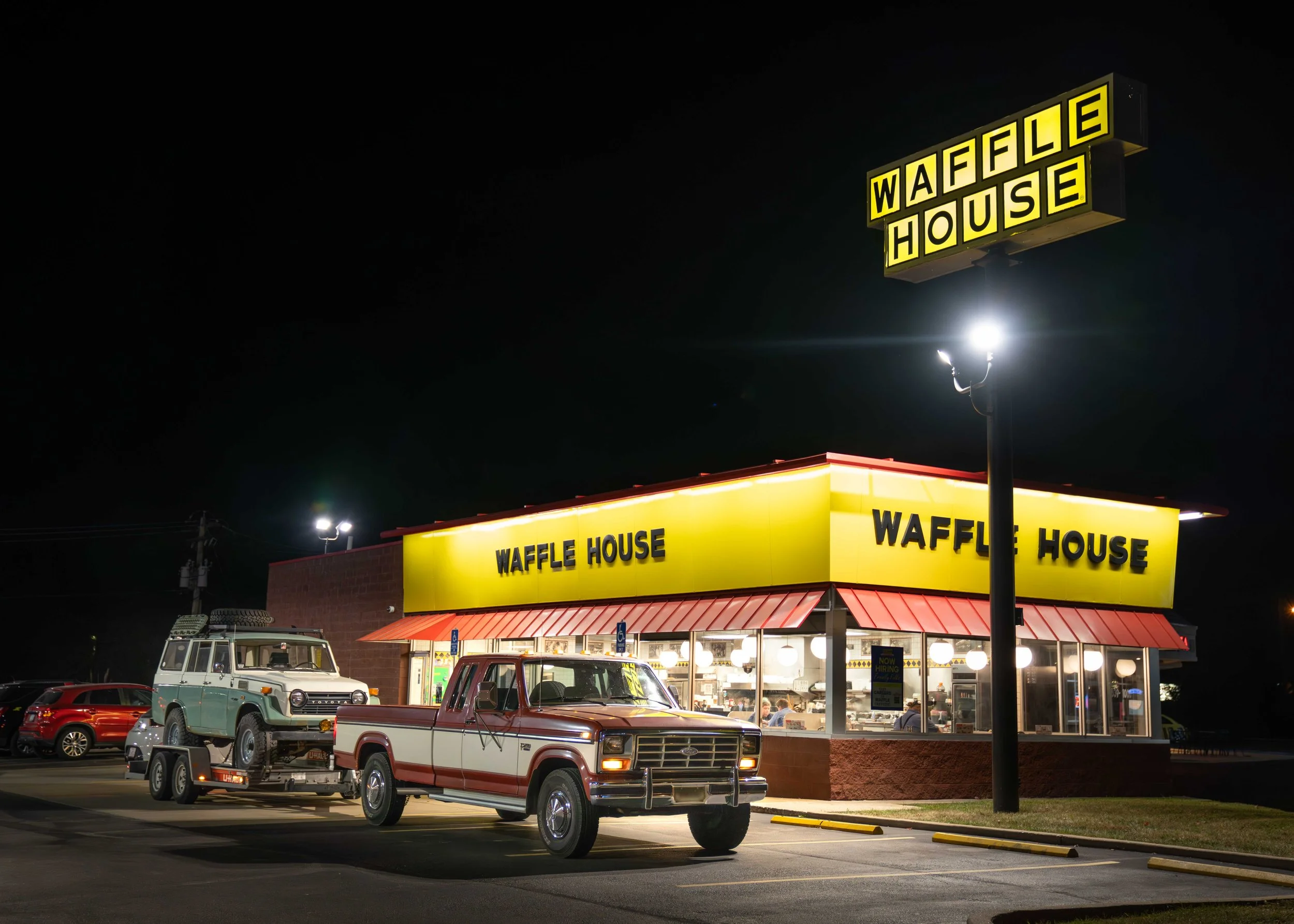 Night view of a Waffle House restaurant illuminated with yellow signage, parked cars in the lot, and a few customers inside.