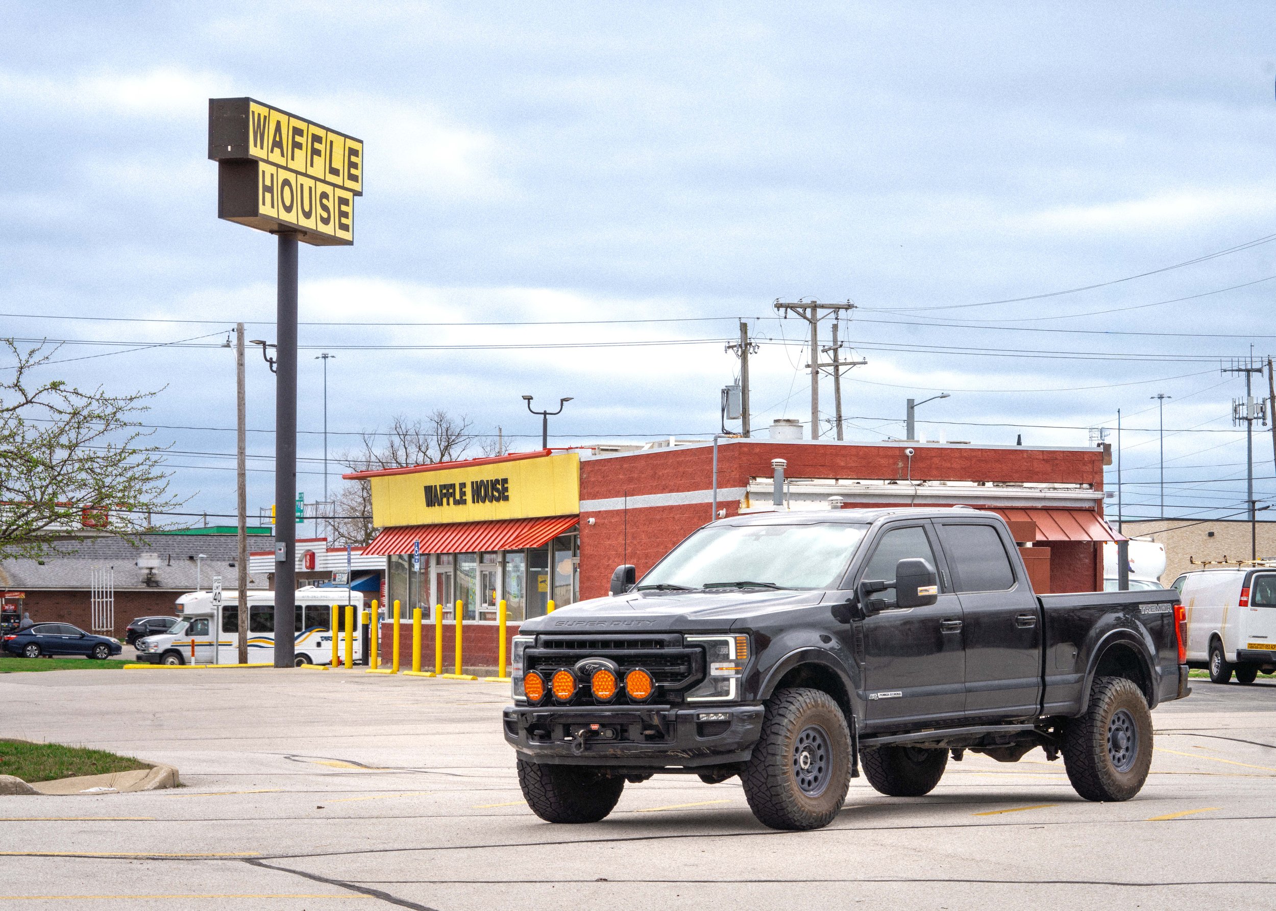 A parking lot in front of a Waffle House restaurant with several cars parked and electric poles in the background under a cloudy sky.