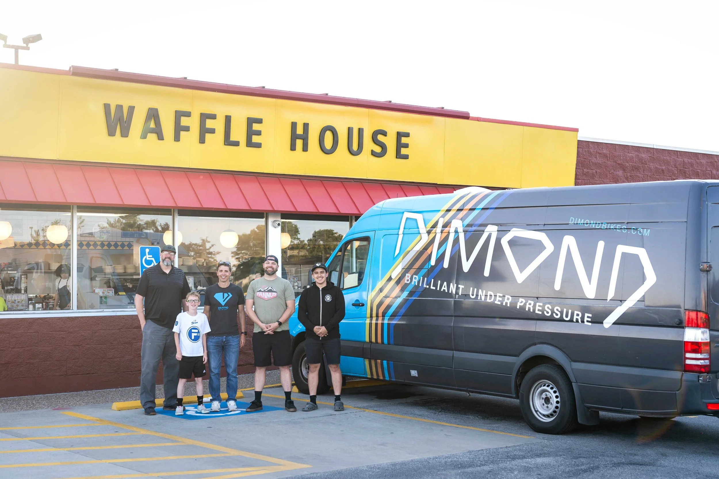 Group of five people standing in front of Waffle House restaurant and a branded van, with customers inside visible through the window.