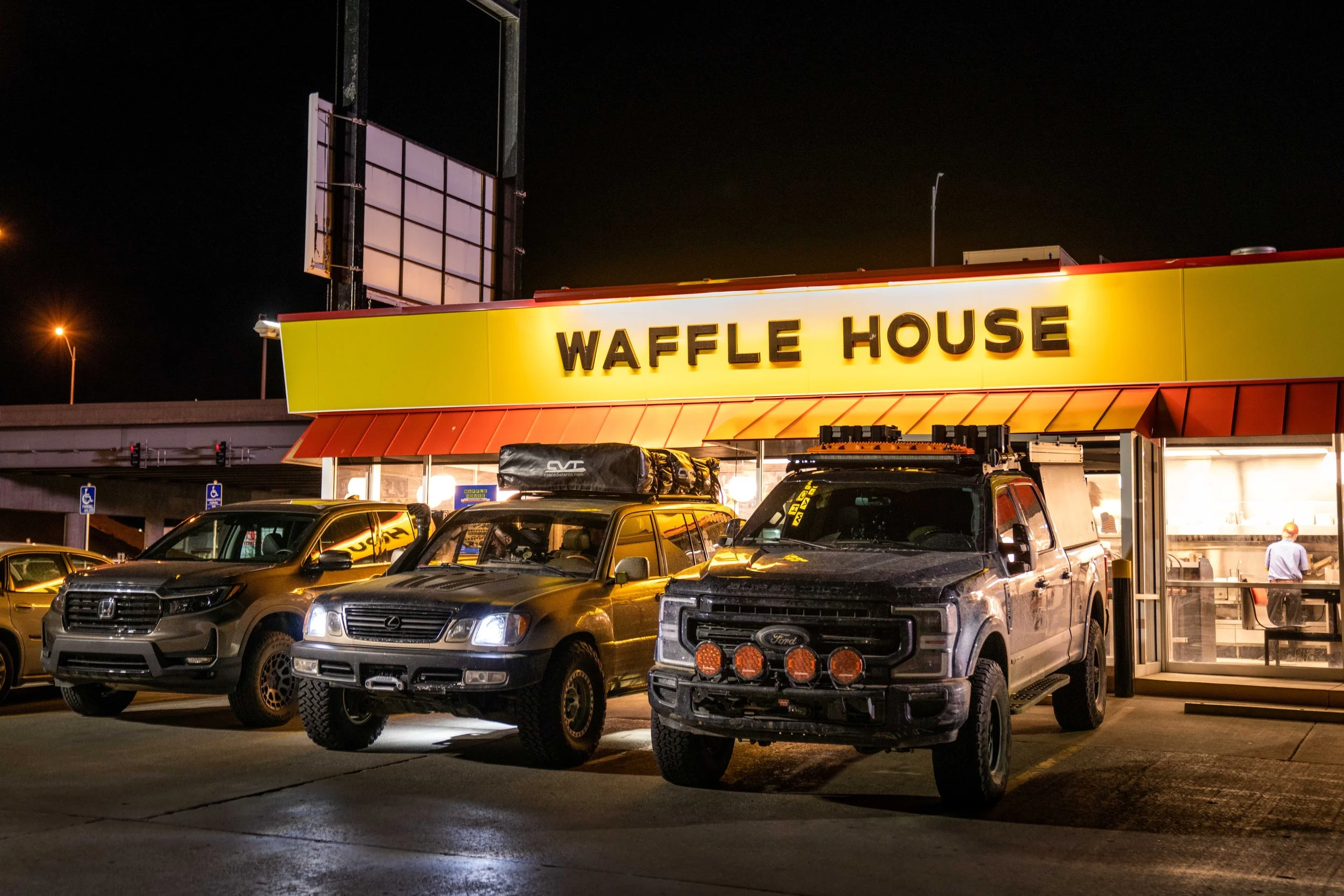 Nighttime view of a Waffle House restaurant with cars parked outside, illuminated signage, and an empty parking lot.