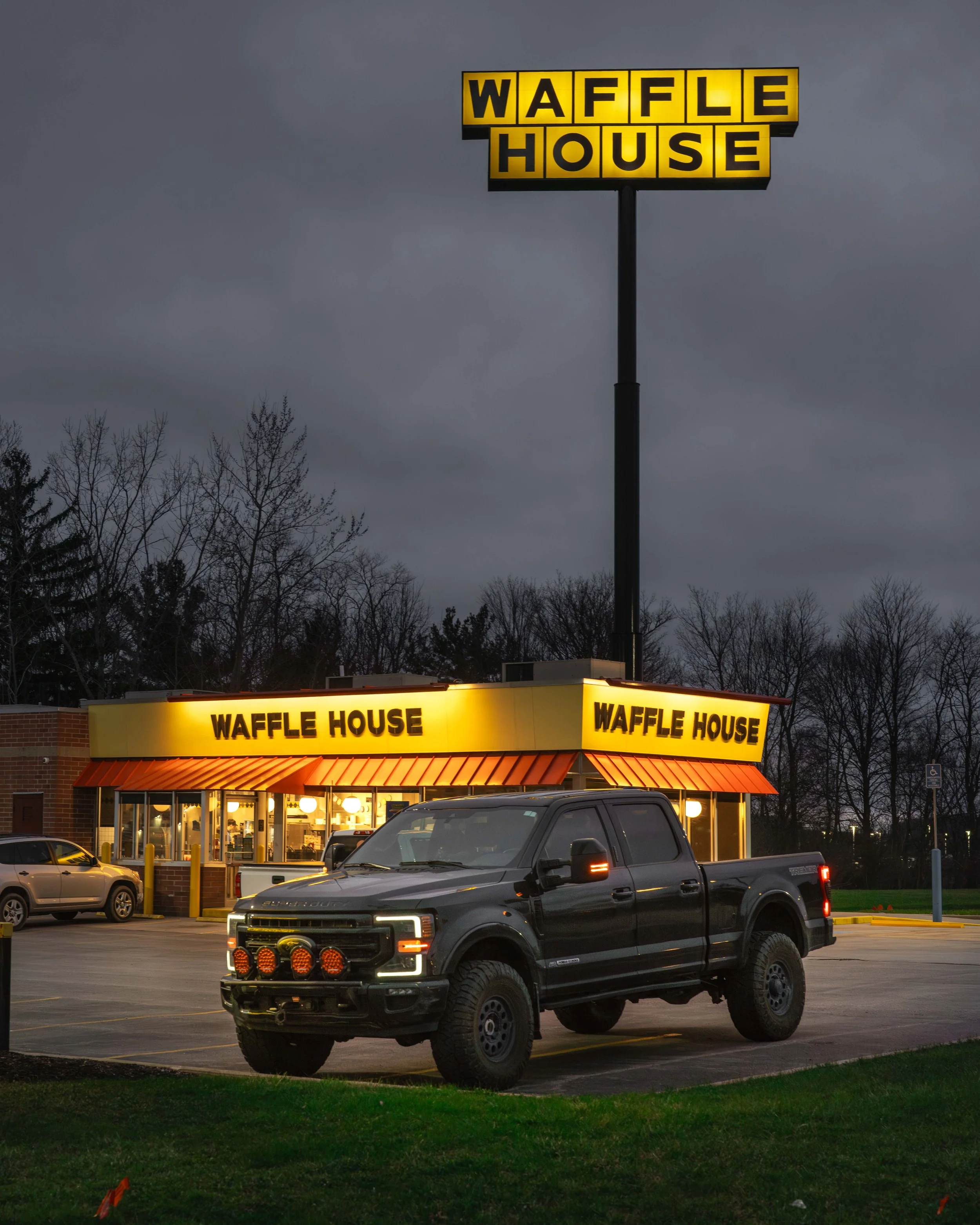 A Waffle House restaurant with its bright yellow illuminated sign and building with a lit interior, cars parked in the parking lot, dark cloudy sky overhead, and leafless trees in the background.