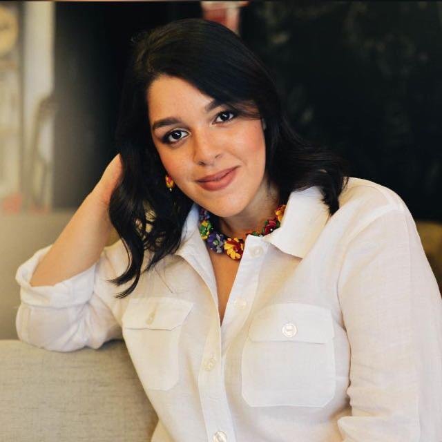 Mujer con cabello oscuro y rizado, vistiendo blusa blanca y collar de colores, sentada en un ambiente interior.