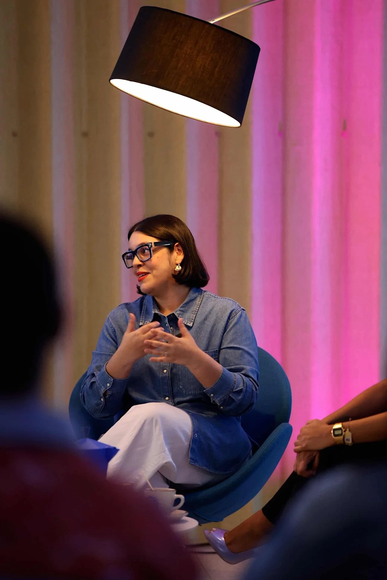Mujer con gafas y cabello corto sentada en una sala, hablando con gestos con otras personas, fondo con cortinas rosa y iluminación cálida.