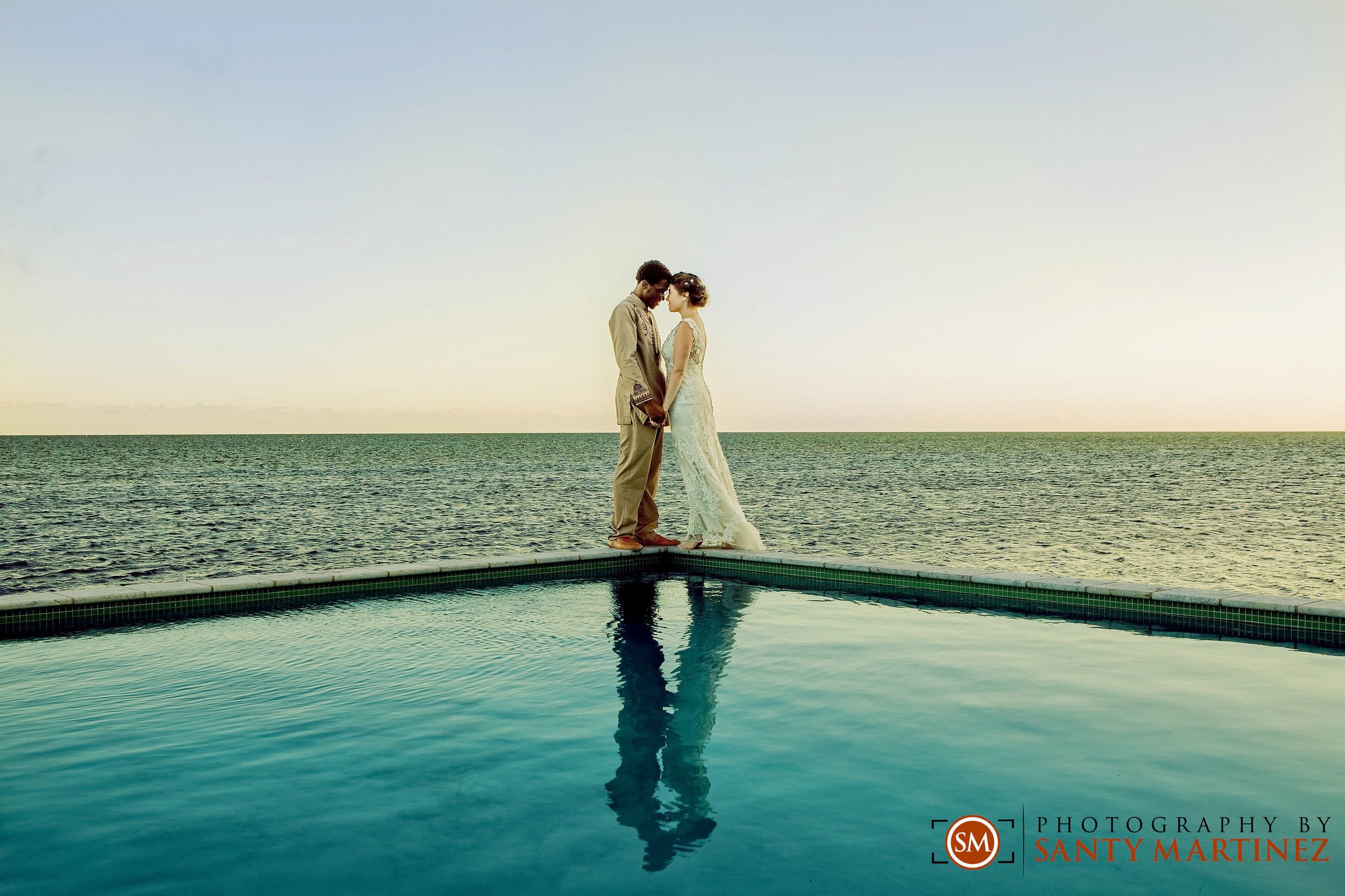 A bride and groom standing at the edge of a pool overlooking the ocean, holding hands and touching foreheads. Their reflection is visible in the water. Photography credit to Santy Martinez.