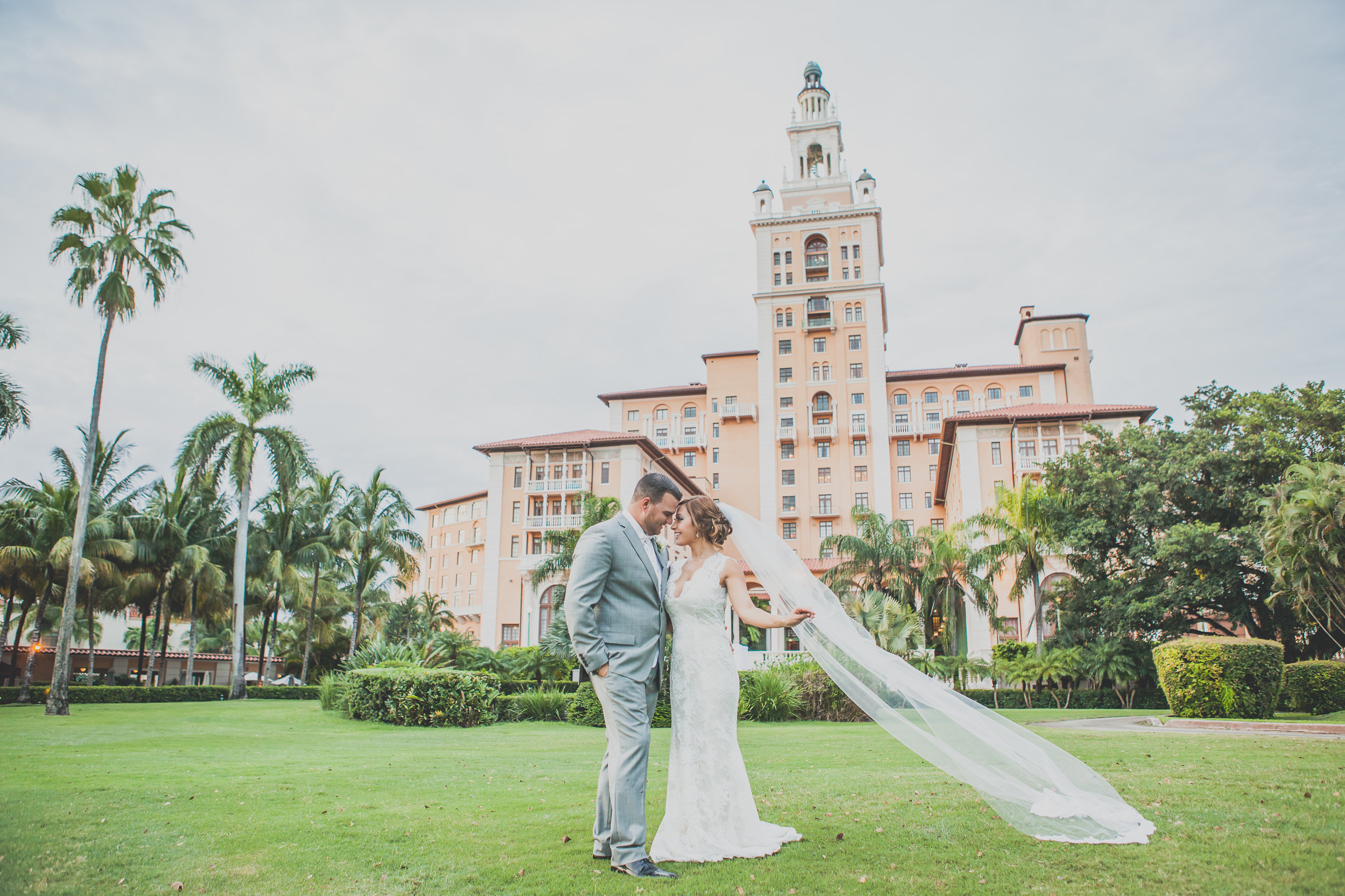 A couple in wedding attire standing on a lawn in front of a vintage-style building with a tower, surrounded by palm trees. The Biltmore Hotel.