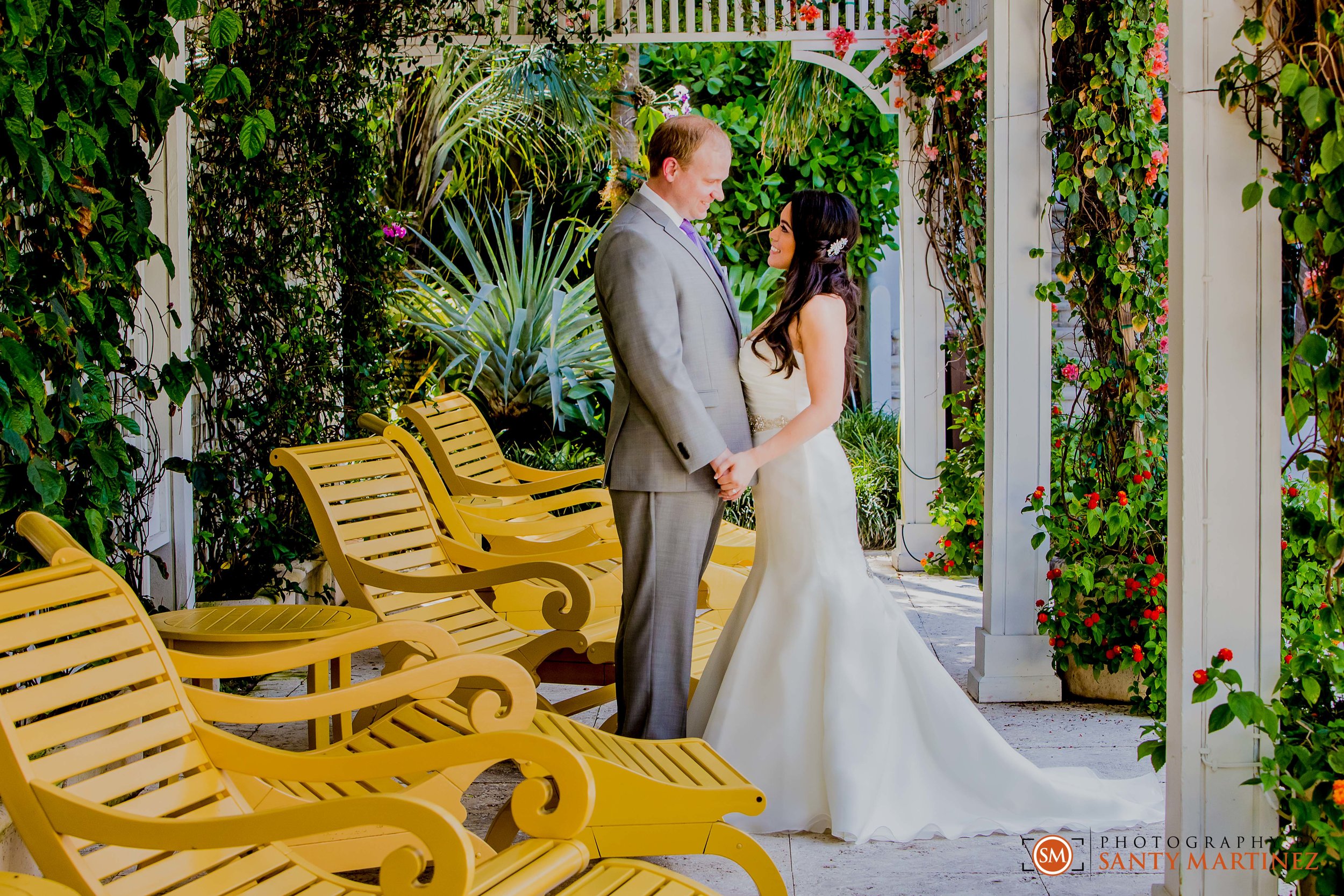 Bride and groom holding hands under a pergola with green foliage and yellow chairs.