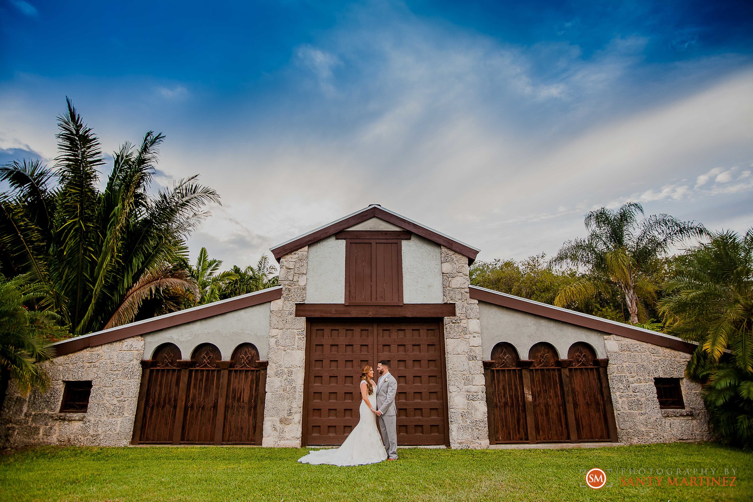 Couple in wedding attire standing in front of a rustic barn with stone walls, large wooden doors, surrounded by palm trees under a blue sky.