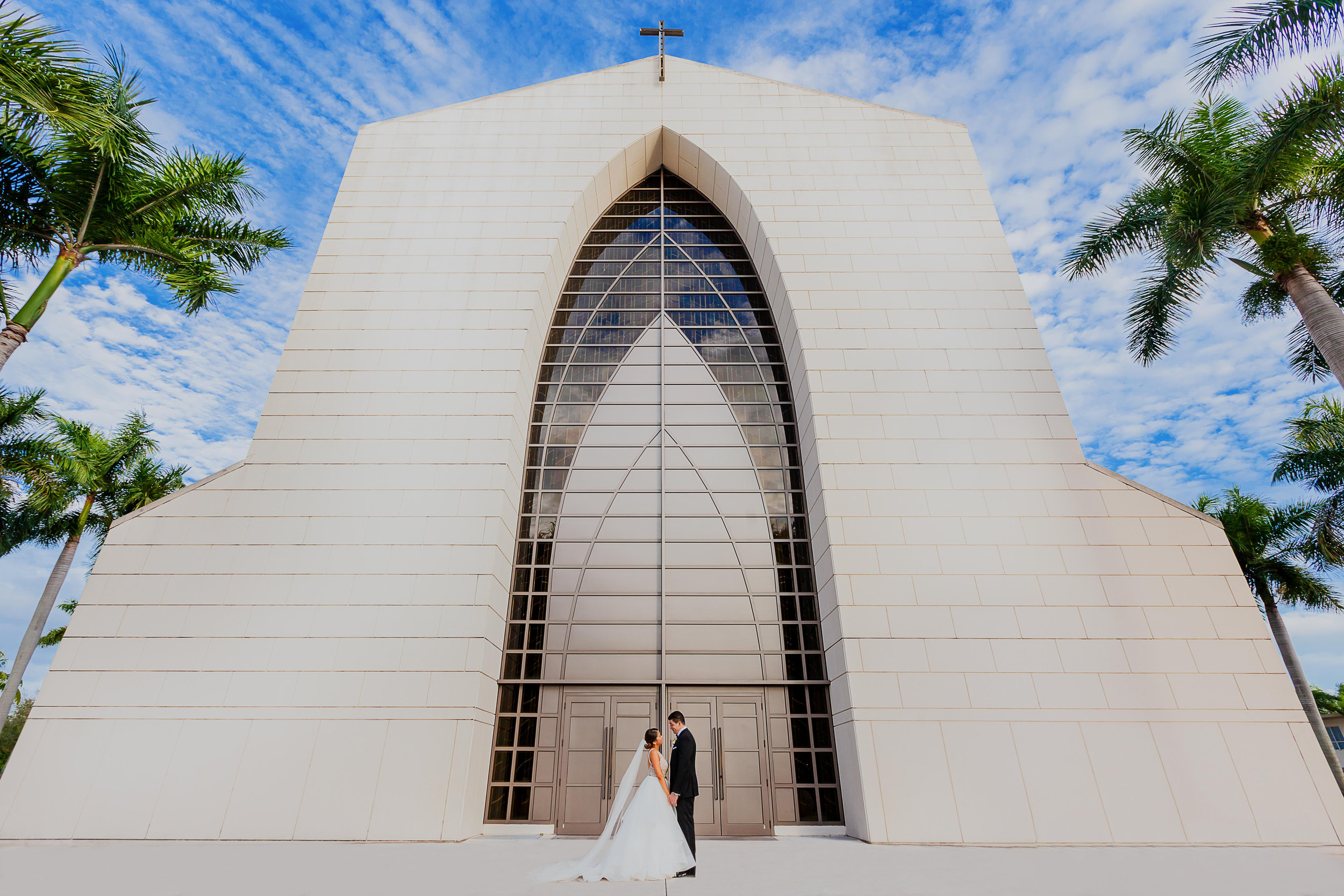 Bride and groom standing in front of a modern church with large arched doorway, surrounded by palm trees, under a blue sky with clouds.