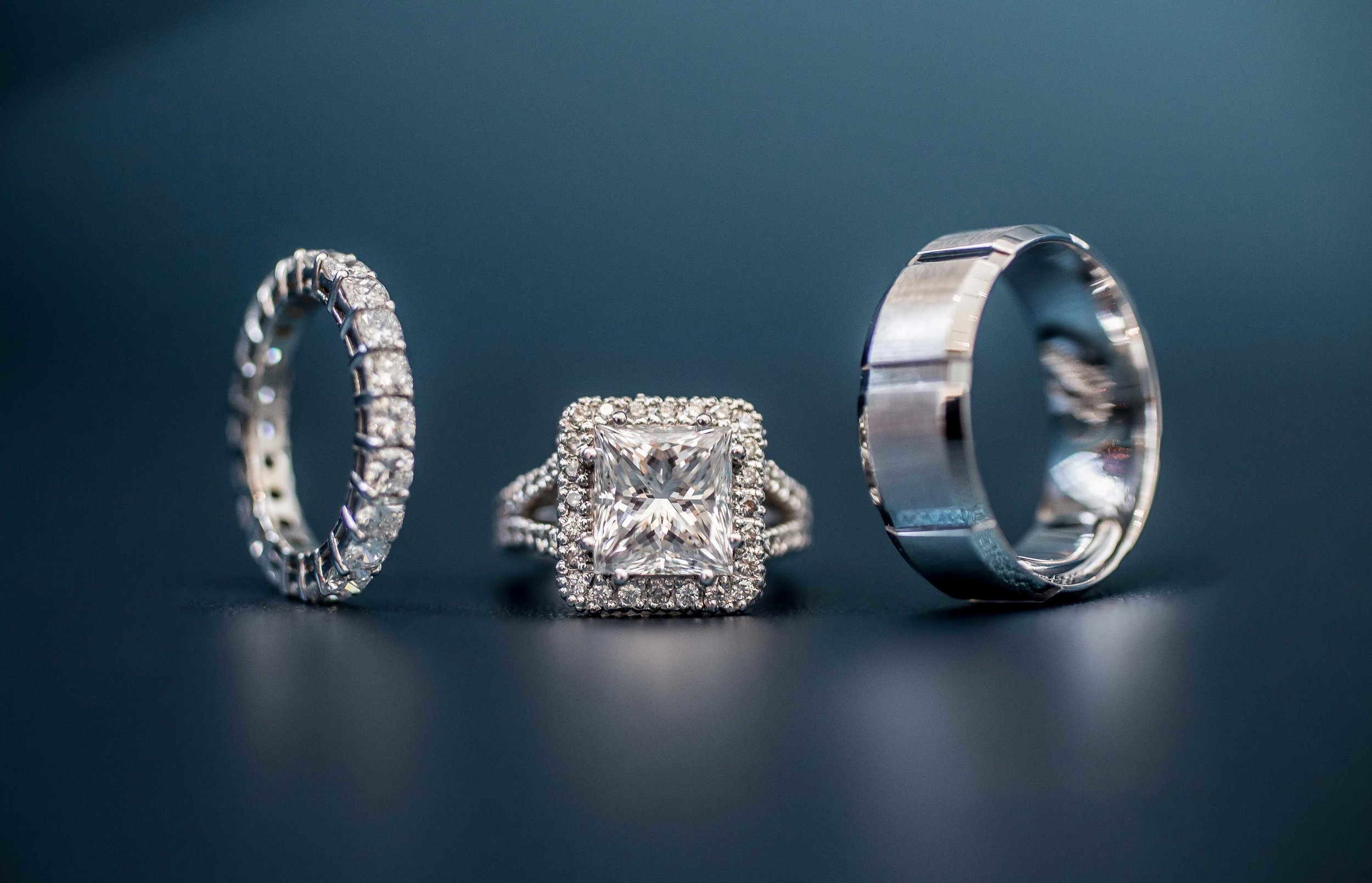 Three rings on a dark surface, including a diamond eternity band, a square-cut diamond engagement ring, and a plain wedding band.