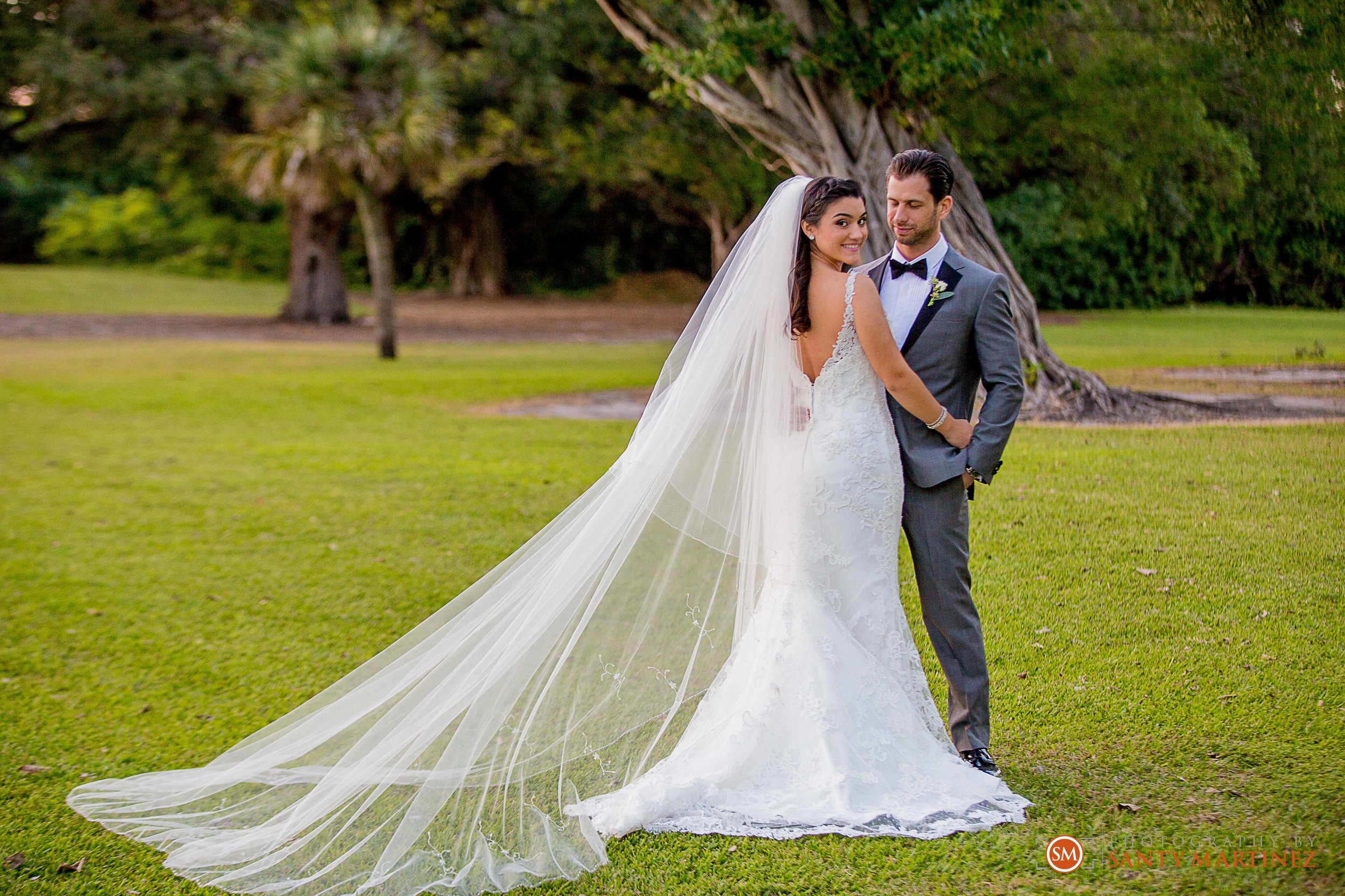 Bride in a white wedding dress with a long veil stands close to a groom in a gray suit with a black bow tie on a grassy area with trees in the background.