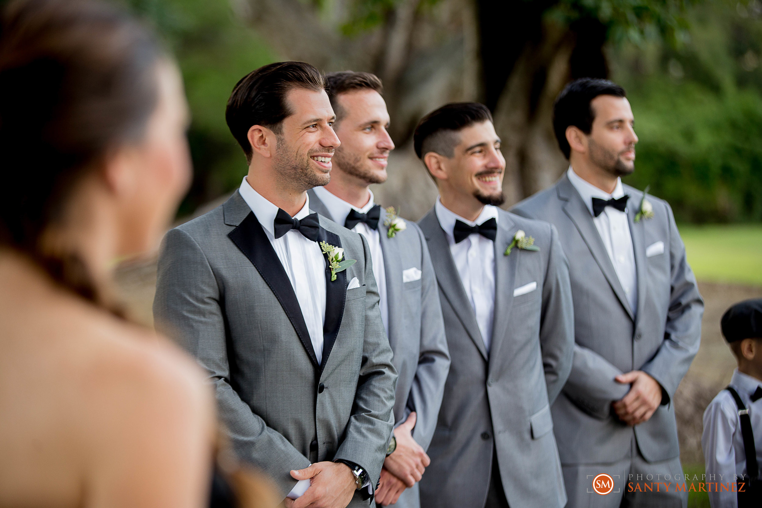 Groomsmen standing in line wearing gray suits and black bow ties at an outdoor wedding.