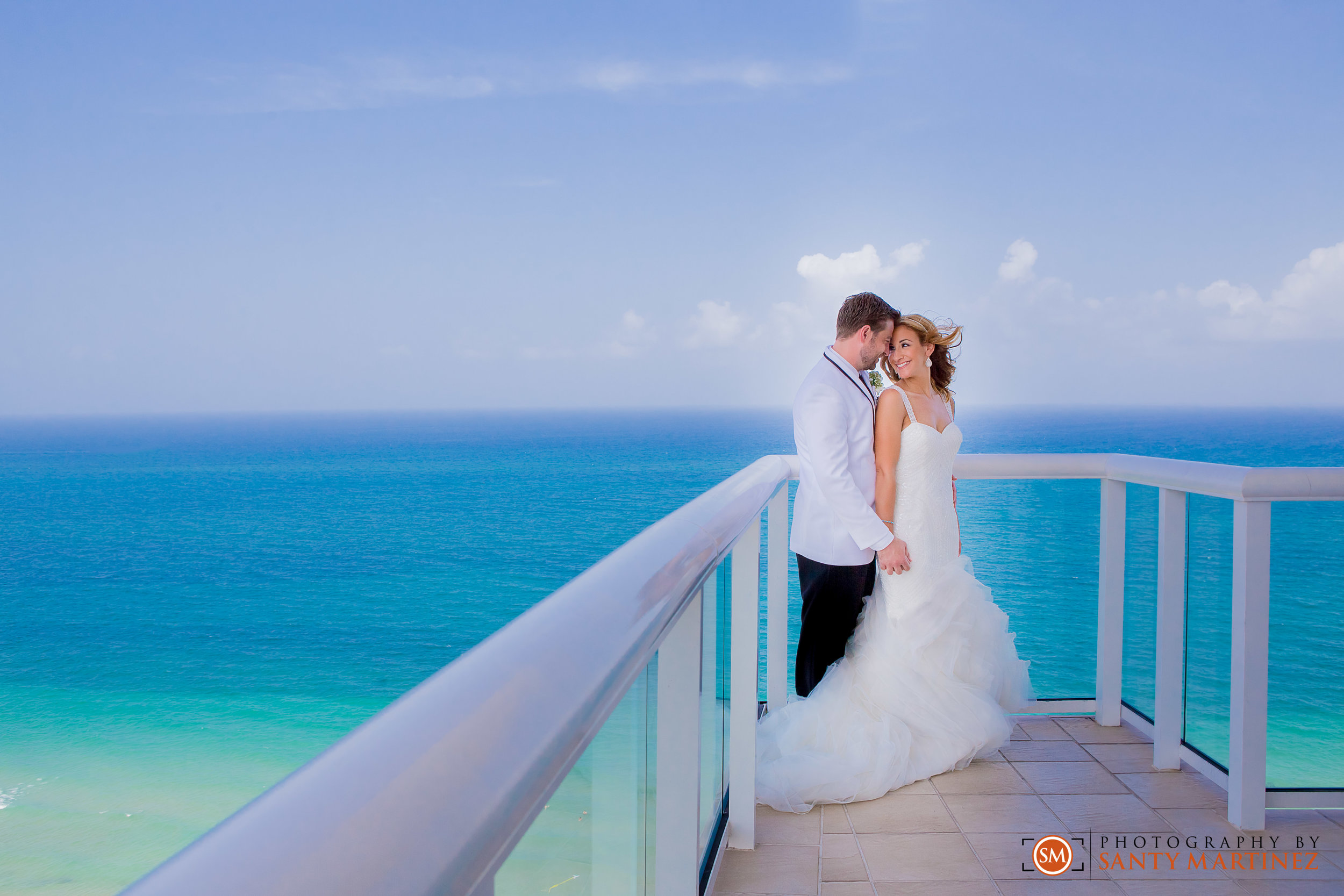 Bride and groom on a balcony overlooking the ocean, with bride wearing a white gown and groom in a white suit.