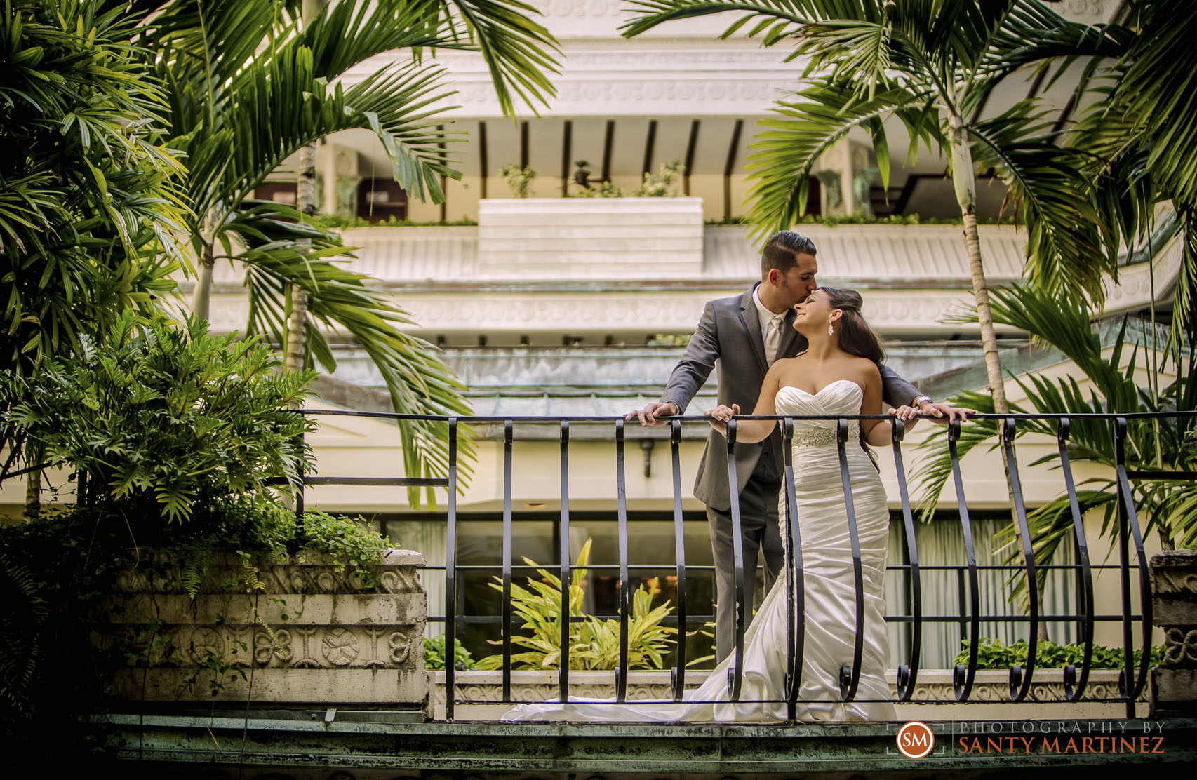A couple posing on a balcony surrounded by lush greenery, with the groom in a gray suit and the bride in a white strapless wedding gown.