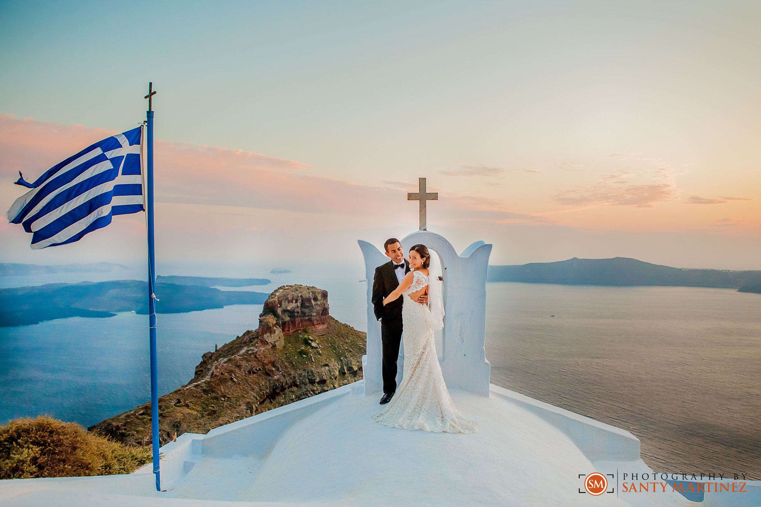 Bride and groom on a white rooftop with a cross in Santorini, Greece, during sunset, with the Greek flag and sea in the background.