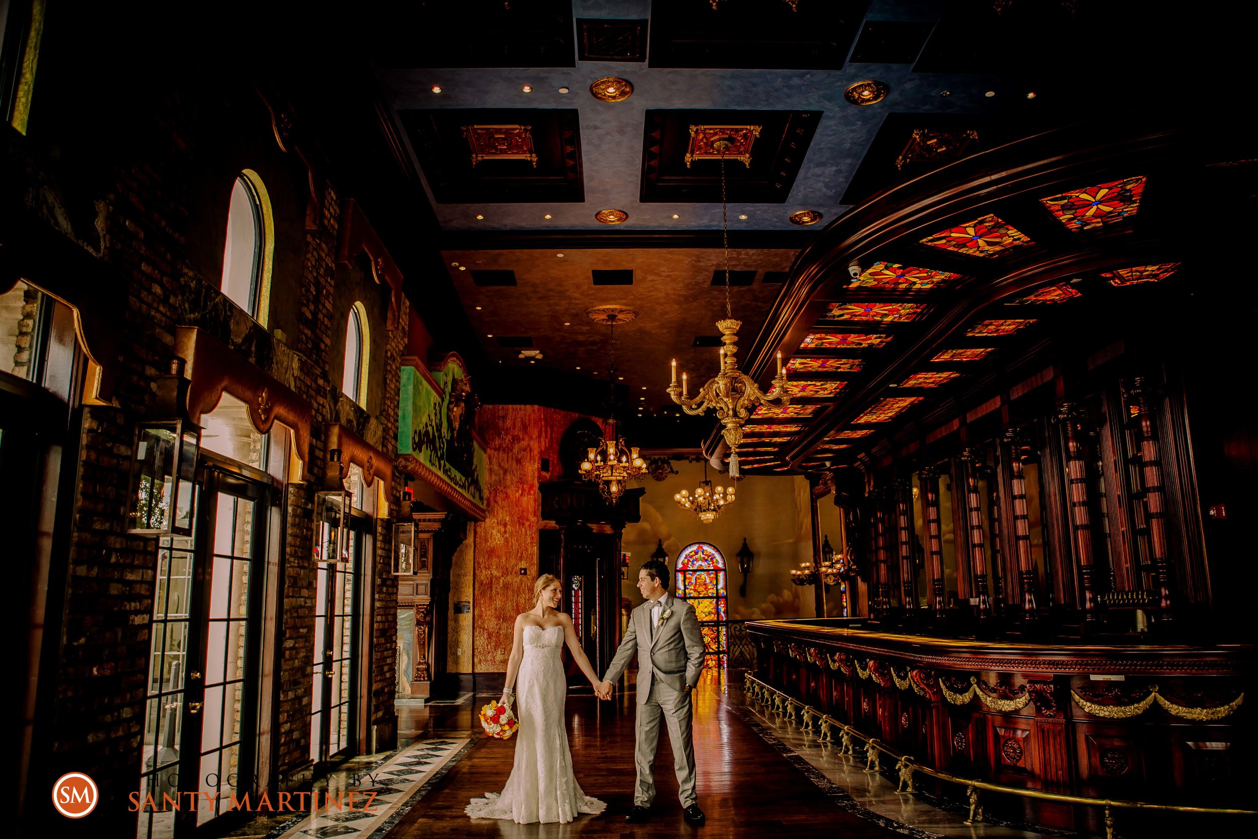 A bride and groom holding hands in an ornate, dimly lit hall with stained glass windows, elaborate chandeliers, and a decorative ceiling.
