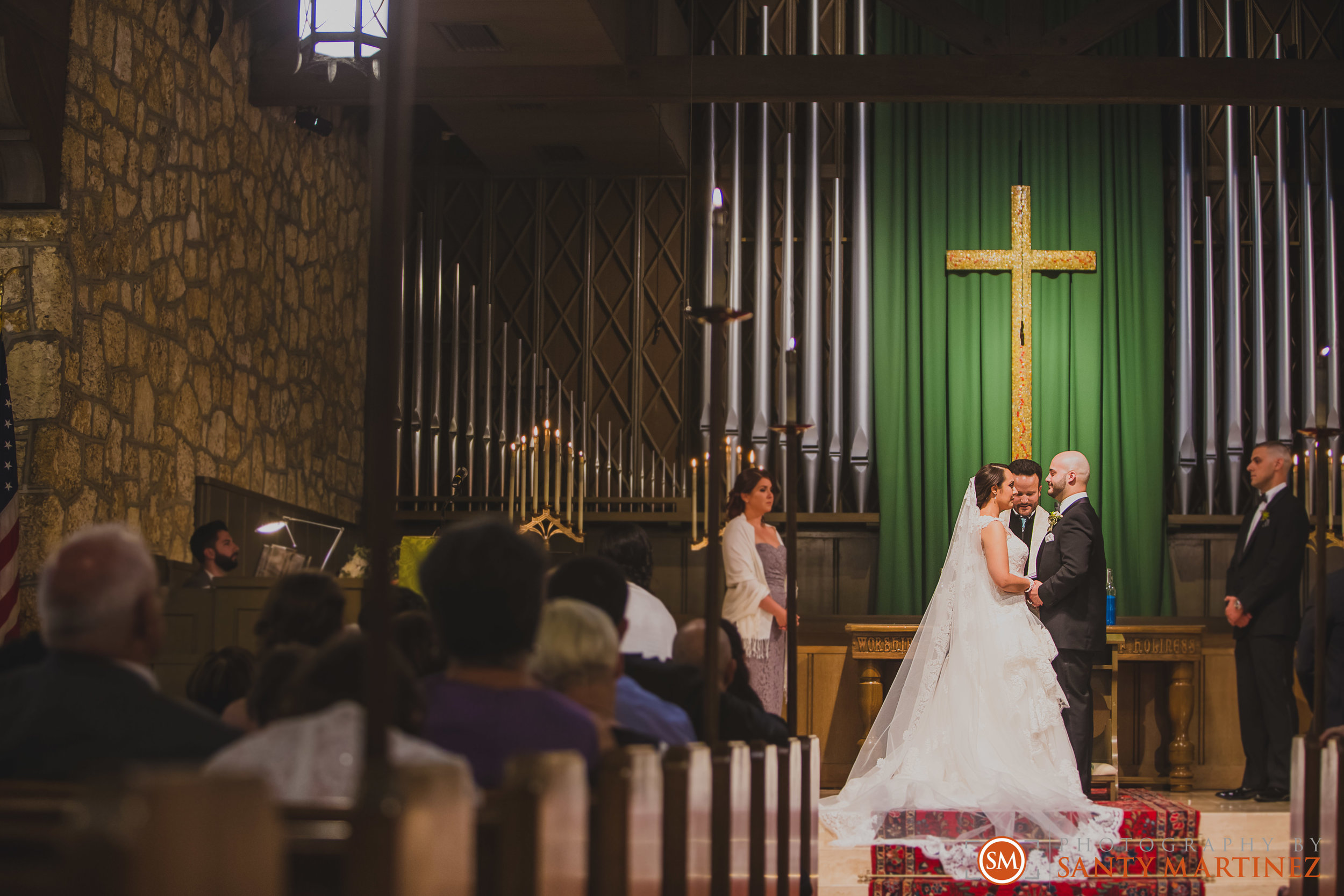 A church wedding ceremony with a bride and groom standing before an altar. A priest is officiating the ceremony. The altar is adorned with a large cross and green curtains. Attendees are seated in pews watching the ceremony.