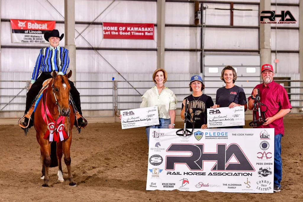 A woman riding a horse with a red ribbon and rosette, standing in an indoor arena, with four people holding oversized checks and a large RHA Rail Horse Association banner in front.