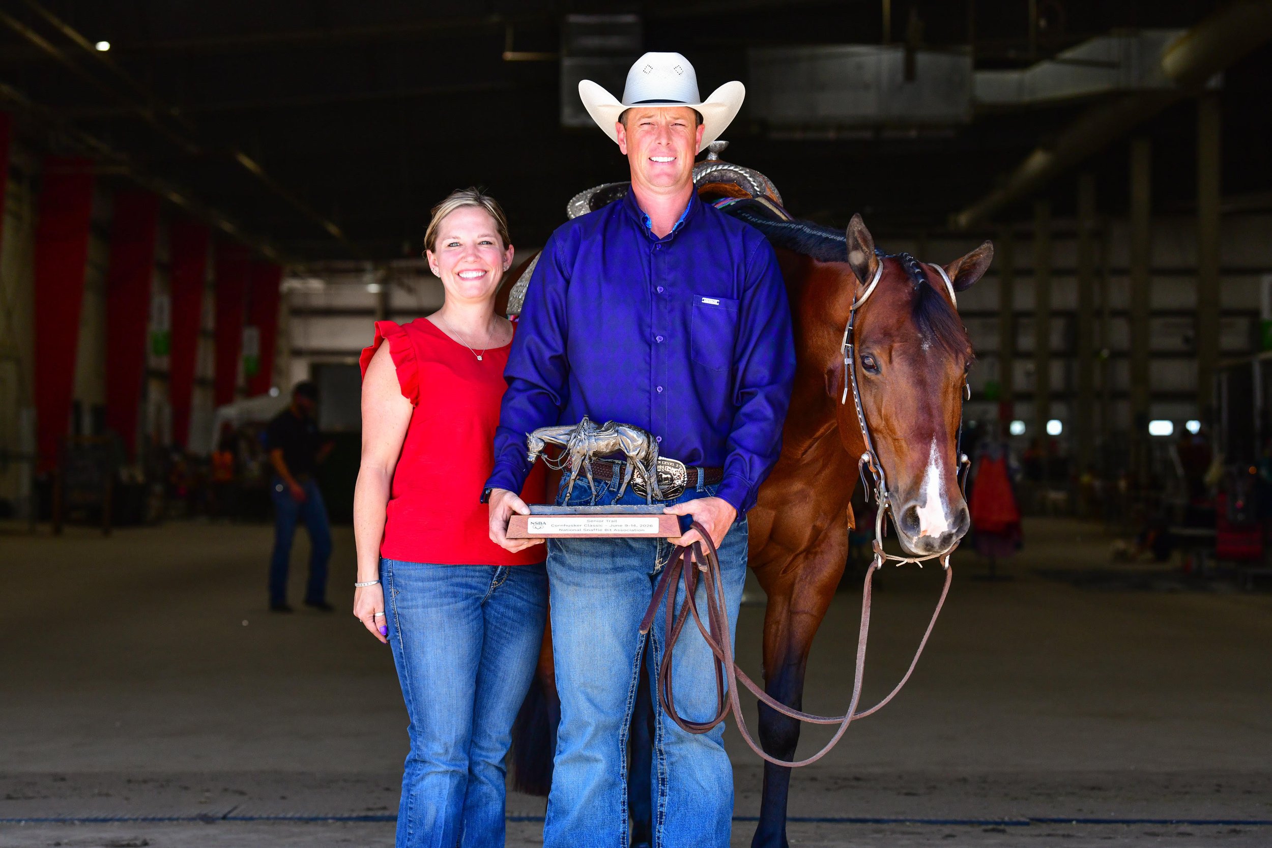 A man wearing a white cowboy hat and a blue long-sleeve shirt standing beside a brown horse with a woman in a red top, inside an indoor arena. The man is holding an award trophy, and the woman is smiling.