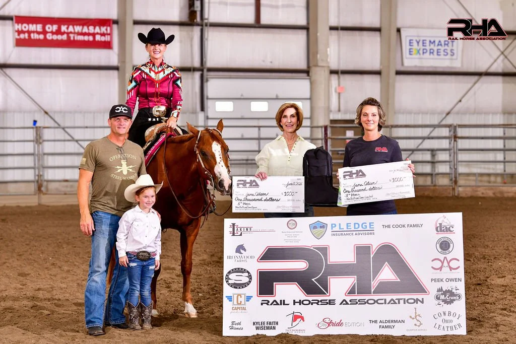Group of five people and a horse in an indoor arena. One woman is sitting on the horse, wearing a Western hat and cowboy boots. Two women and two children are standing in front, holding oversized checks and a trophy. A large banner with the Rail Horse Association logo is displayed in front of them.