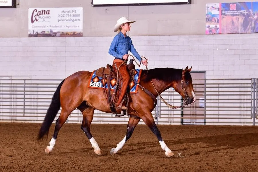 A woman wearing a cowboy hat and blue shirt riding a brown horse inside an indoor arena.