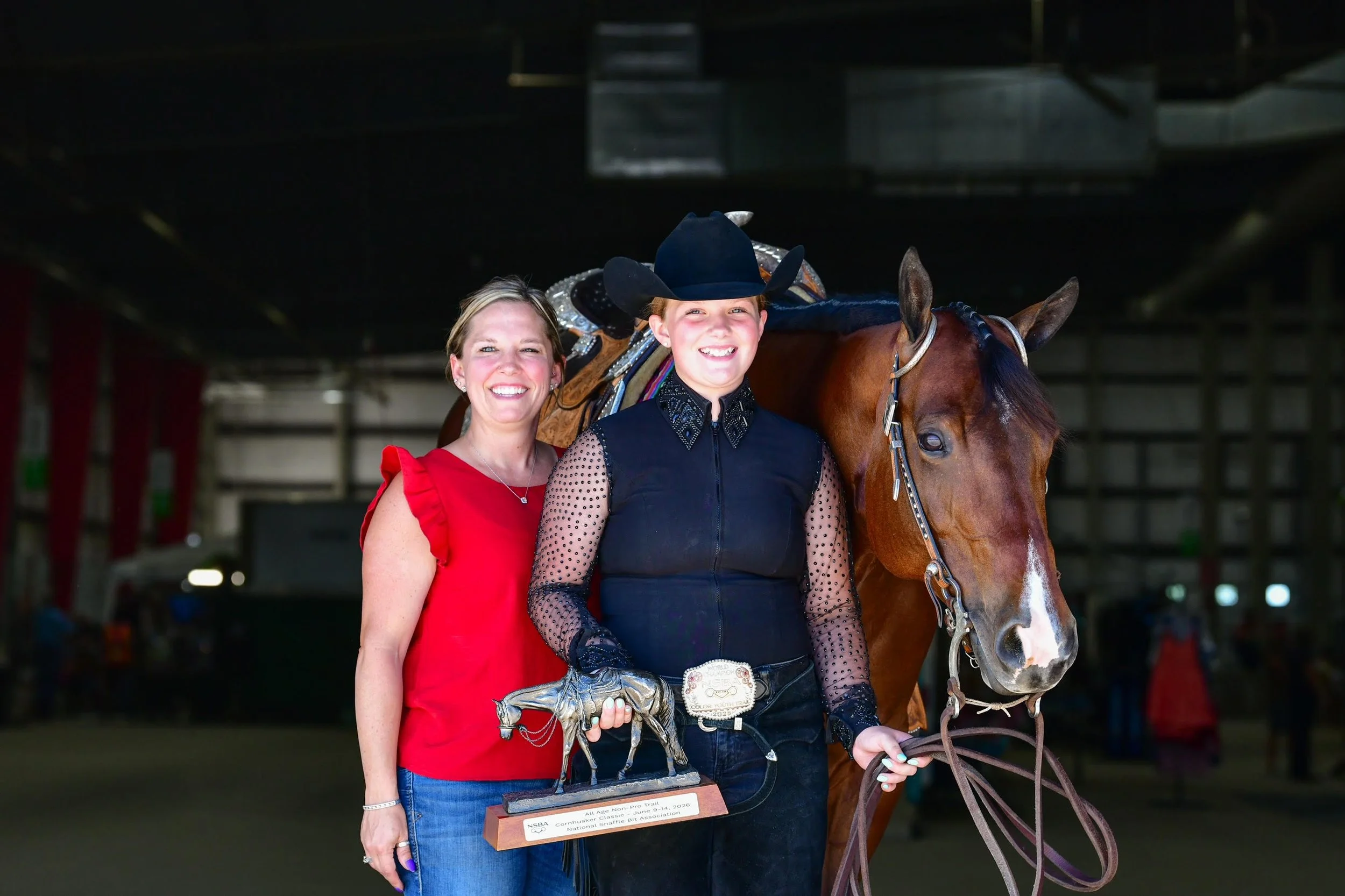 A woman in a red dress and a young person in a black cowboy hat and black lace top standing next to a brown horse, holding a trophy, inside a dimly lit stable or indoor equestrian arena.