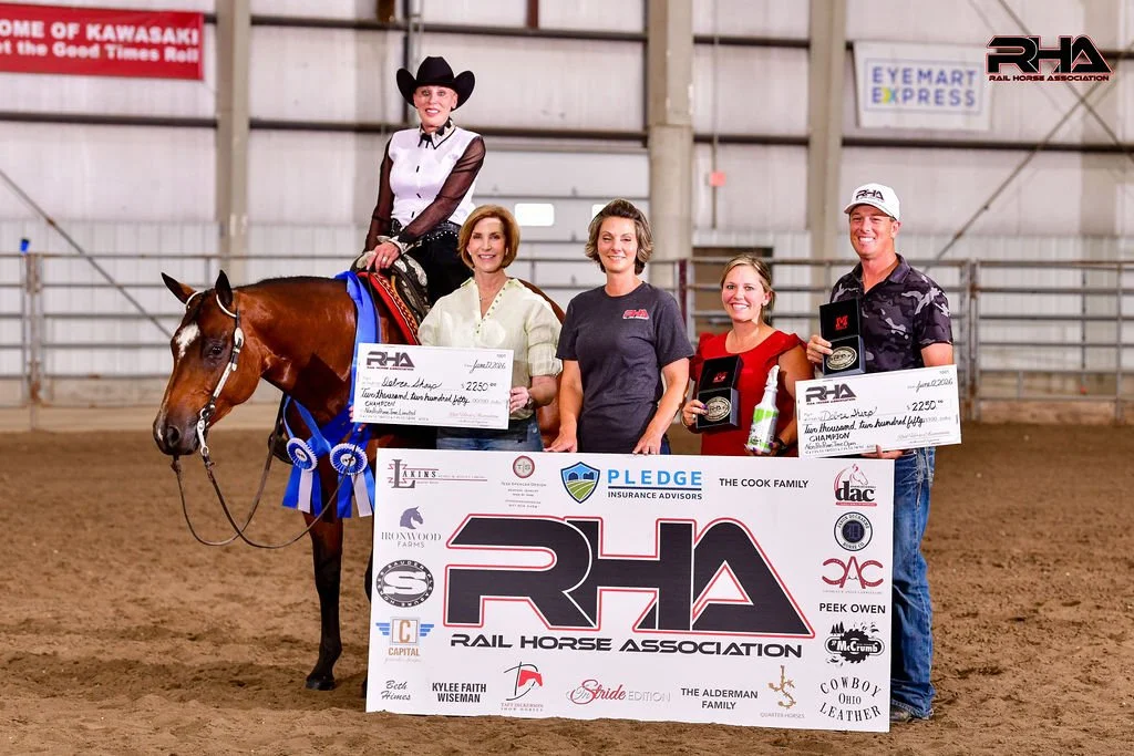 A woman dressed in Western riding attire sitting on a brown horse with blue ribbons at an indoor equestrian event. Four people stand beside her, holding large checks, plaques, and awards. A large RHA Rail Horse Association banner displays sponsor logos and the event details in front of them.