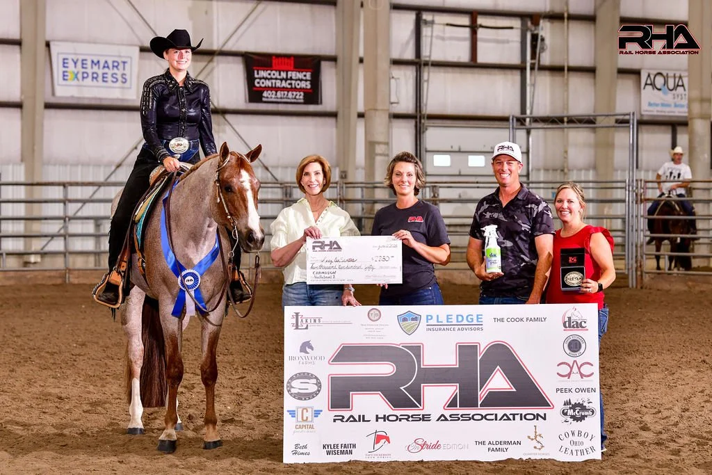 A woman on horseback wearing a black cowboy hat and black outfit, holding a ribbon. Four other women are standing nearby, holding a large check, a bottle of cleaning spray, and an award plaque, inside a rodeo arena. There are banners and signs on the walls.