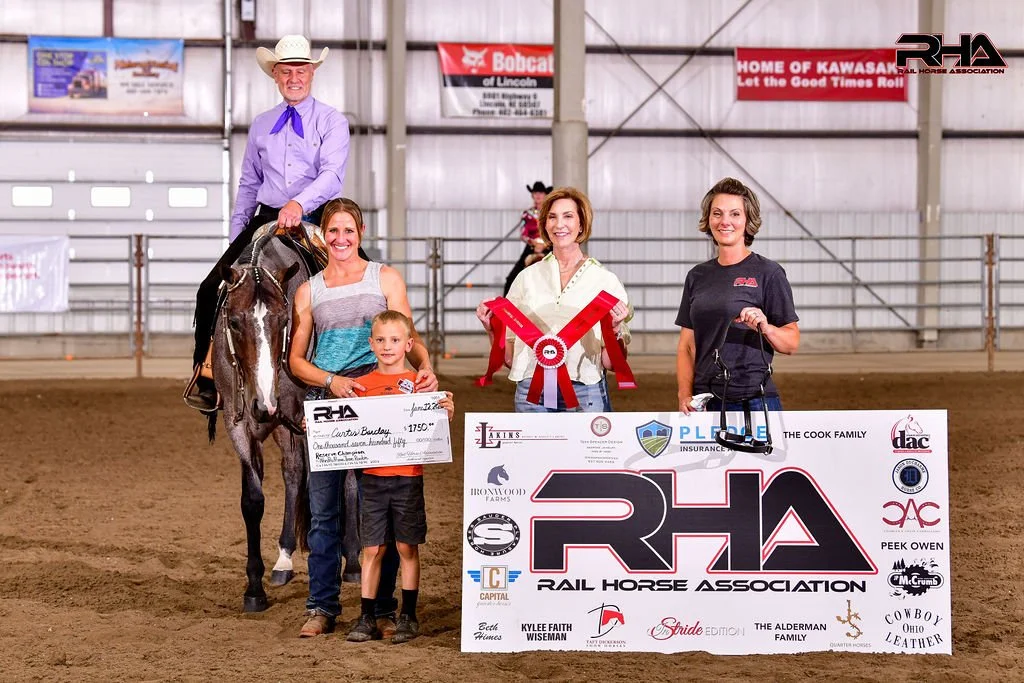 Group of people at a rodeo or equestrian event, holding a large check, a large ribbon, and a mini horse, inside an indoor arena with banners in the background.