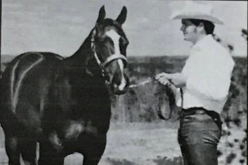 A woman in a wide-brimmed hat and white shirt standing outdoors, holding a horse by the reins.