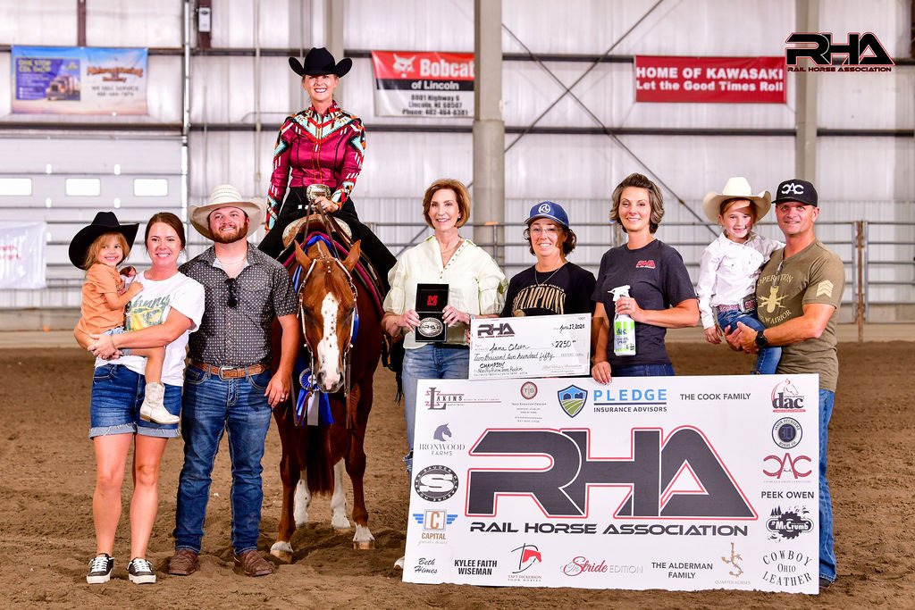 Group of ten people, including children, standing inside a rodeo arena with a horse and rider, celebrating a championship win. They are holding a large check, a plaque, and medals, with banners and advertisements hanging on the wall behind them.