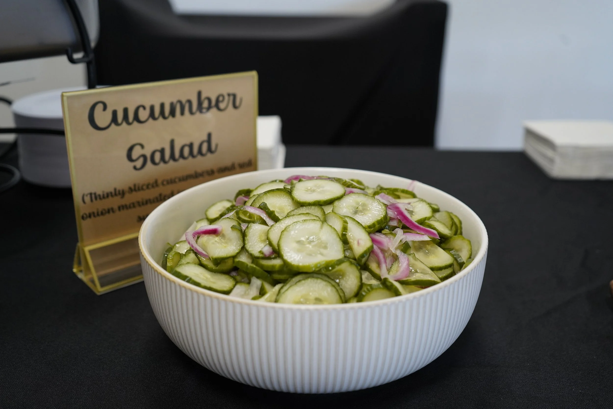 Bowl of sliced cucumbers with red onions on a black table, labeled 'Cucumber Salad' in the background