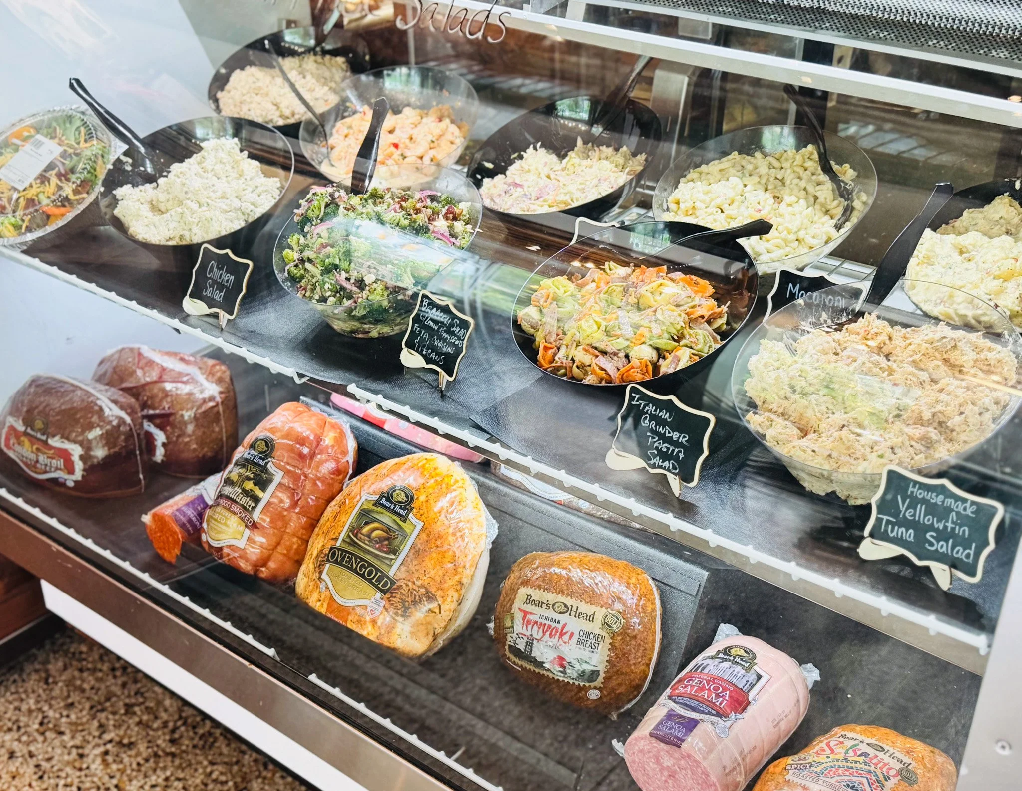 Deli display case with large hams, cheese, and various meats, decorative lemons, and greenery inside, with pack of pre-packaged meats and cheeses on top.