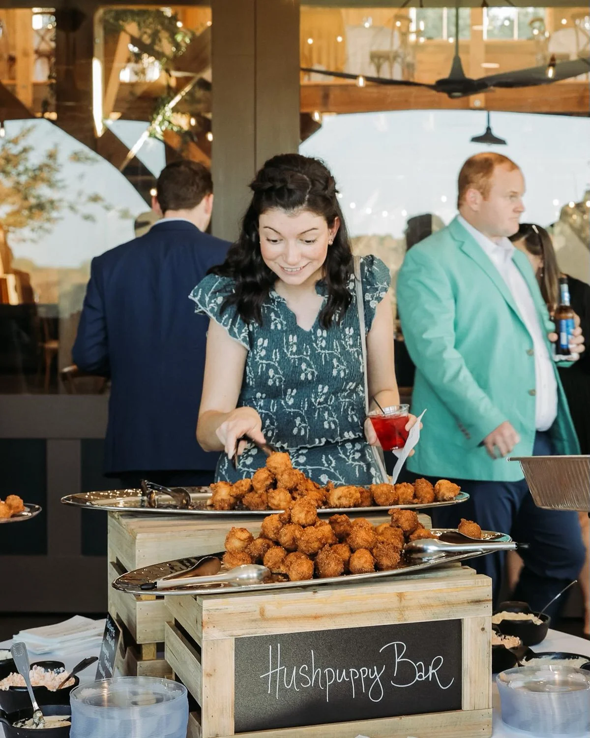 Woman serving fried food at a bar labeled 'Hushpuppy Bar' in an indoor/outdoor setting during daylight.