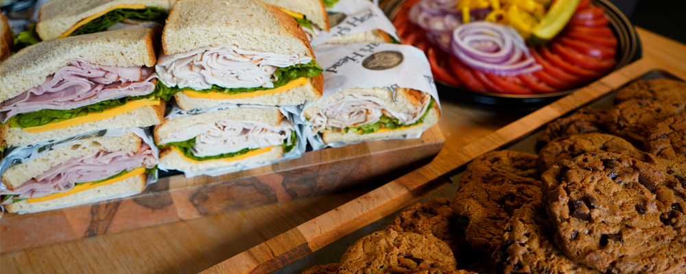 Sandwich platters from the deli with turkey sandwiches with lettuce and cheese on white bread, a bowl of sliced tomatoes and onions, and several chocolate chip cookies.