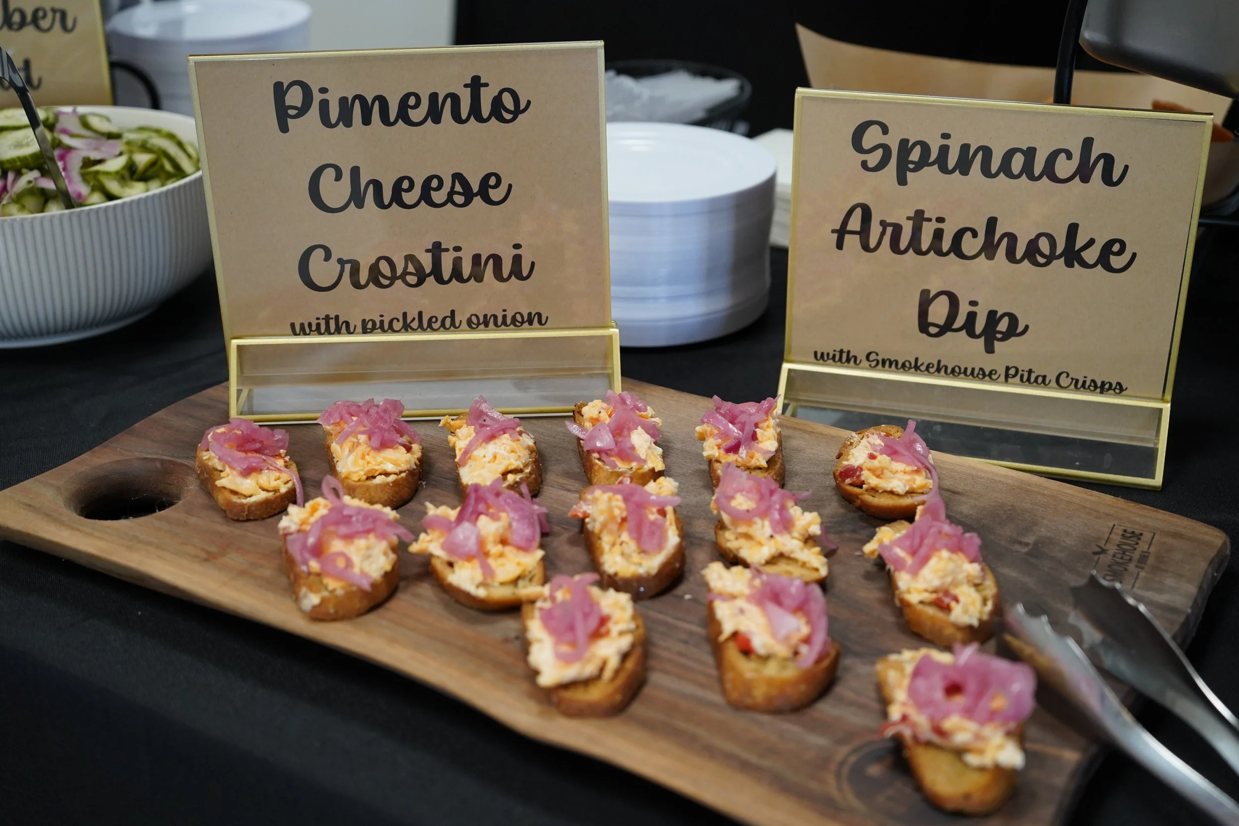 Appetizer bites with scrambled eggs and pink pickled onions on toasted baguette slices, served on a wooden tray at a buffet, with signs labeled "Pimento Cheese Crostini with pickled onion" and "Spinach Artichoke Dip with Smokehouse Pita Crisps."