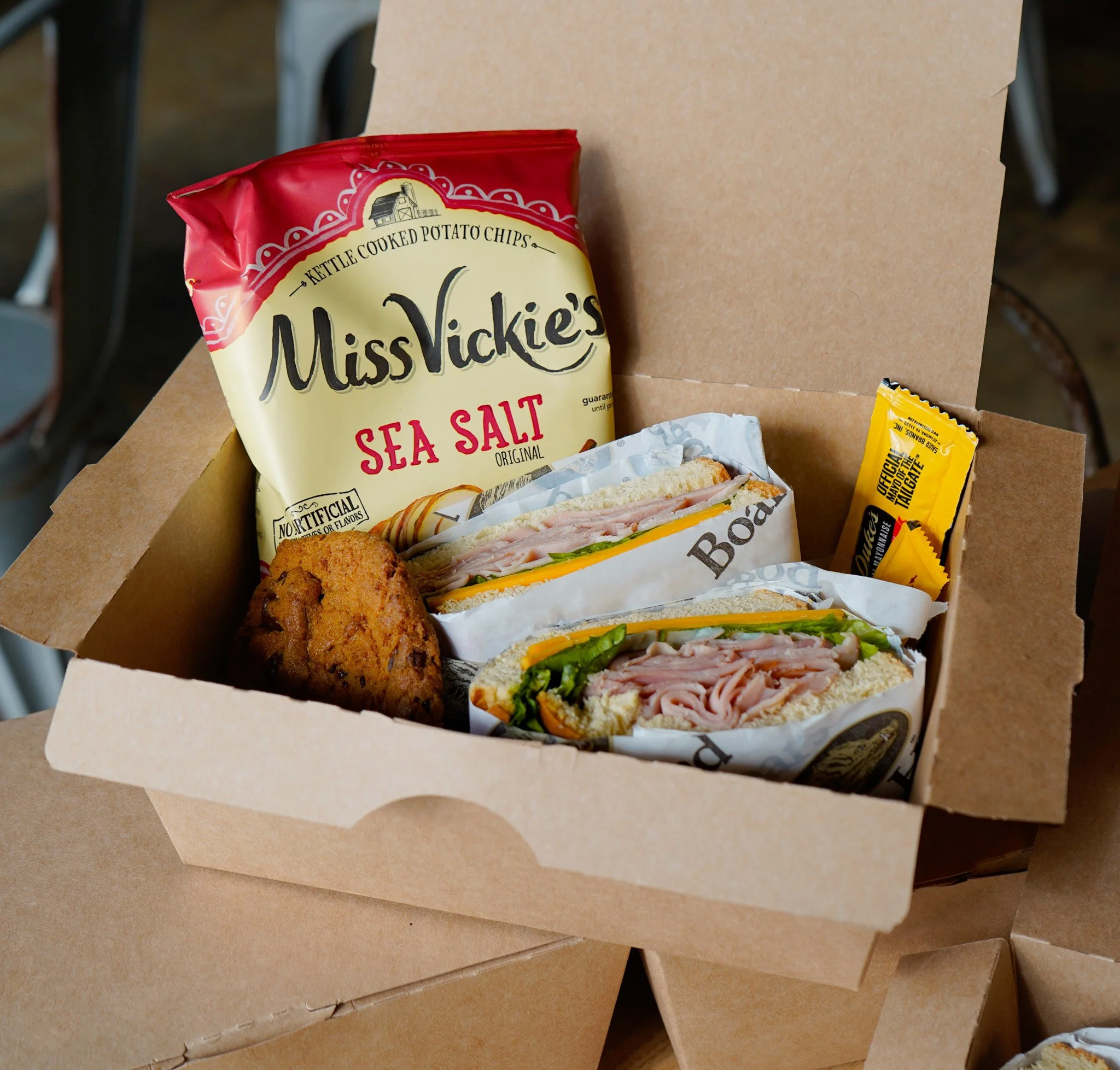 Boxed lunch including a whole turkey sandwich with cheese and lettuce on white bread, a chocolate chip cookie, your choice of side, cutlery, mustard, and mayo.