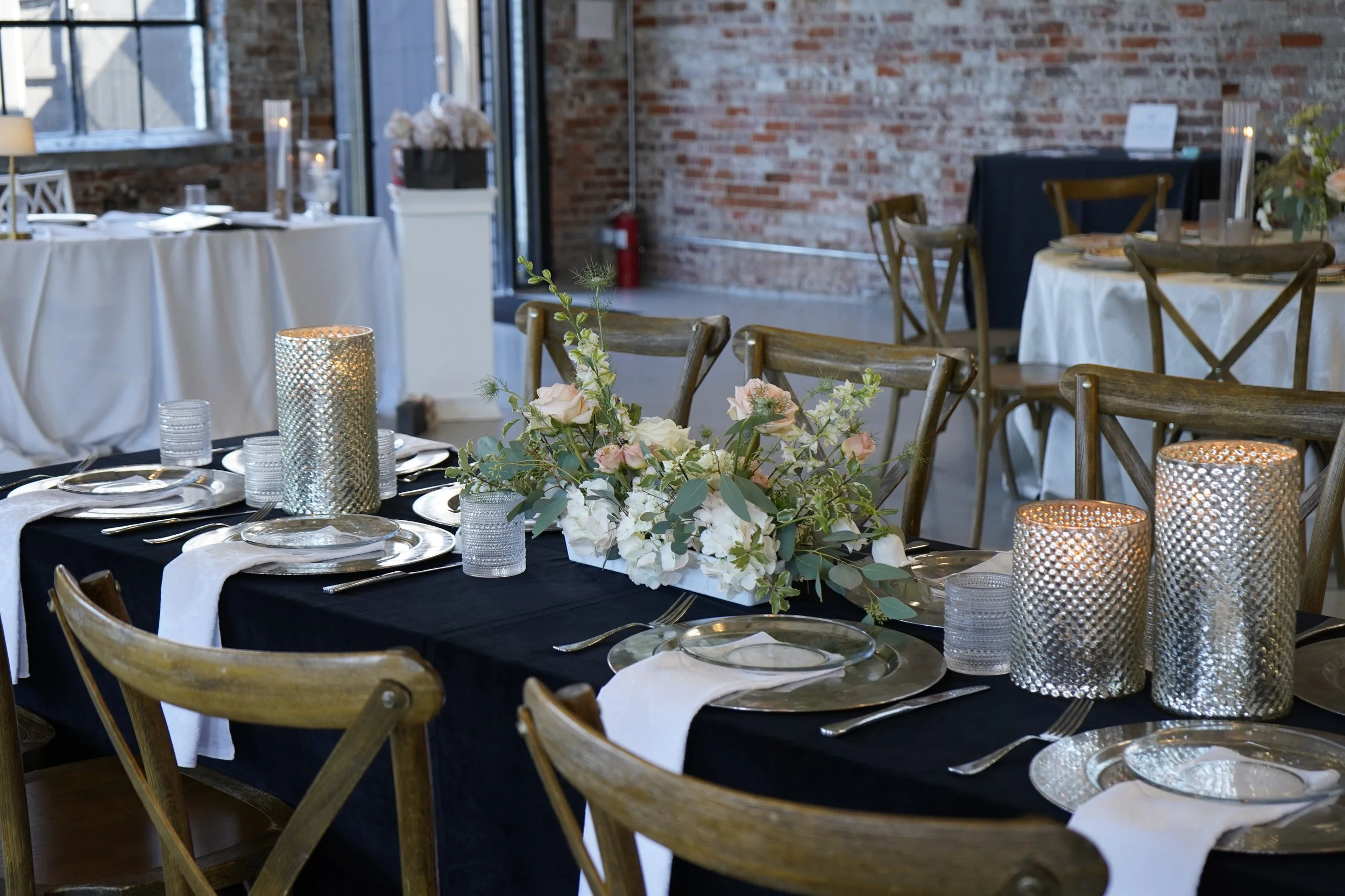 Elegant dining table set for a formal event with floral centerpiece, candles, and place settings in a rustic venue with exposed brick walls.