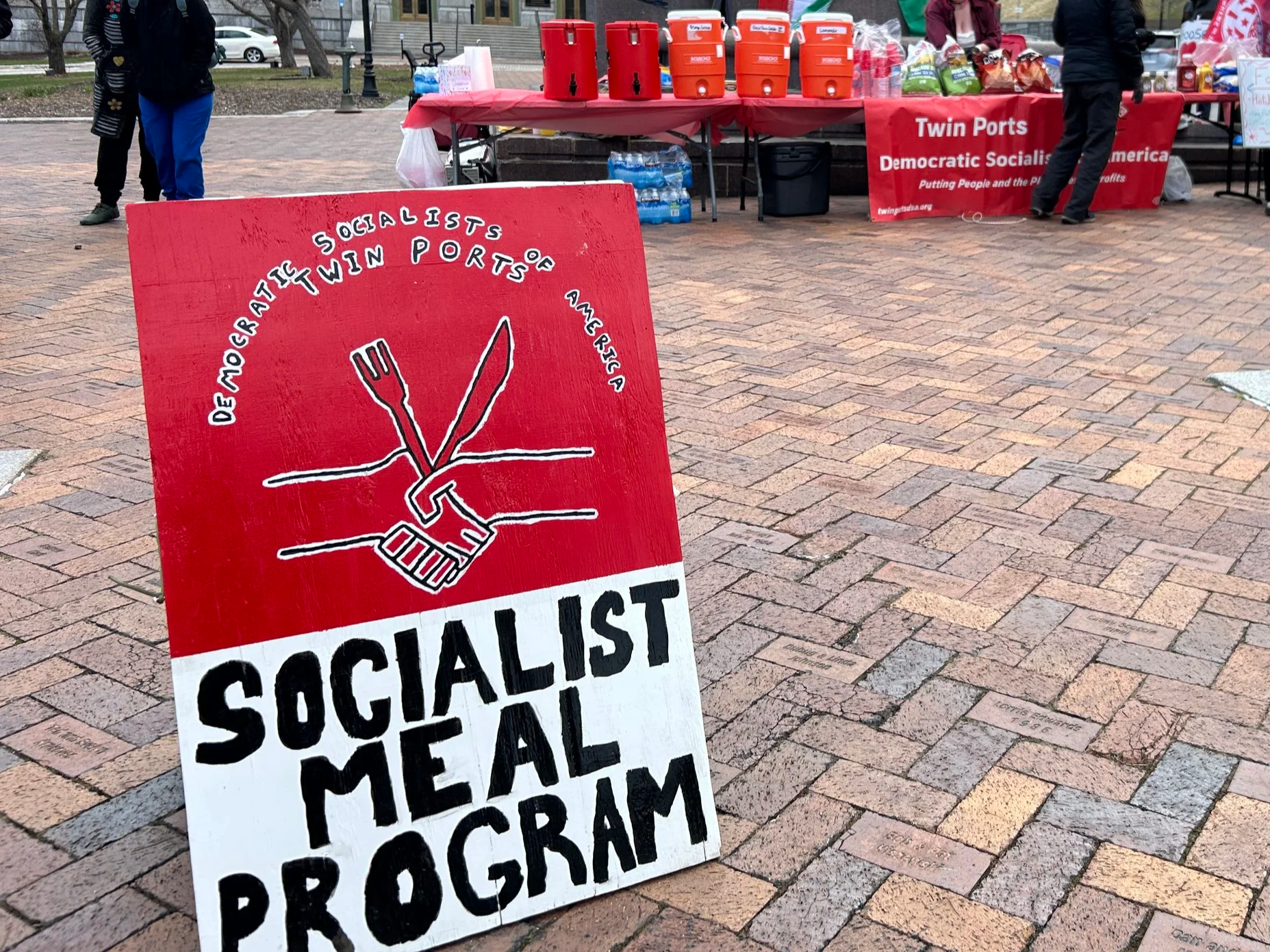 A-frame sign sits out on brick plaza. Sign reads Socialist Meal Program with a red logo showing two hands shaking while clasping a fork and knife.