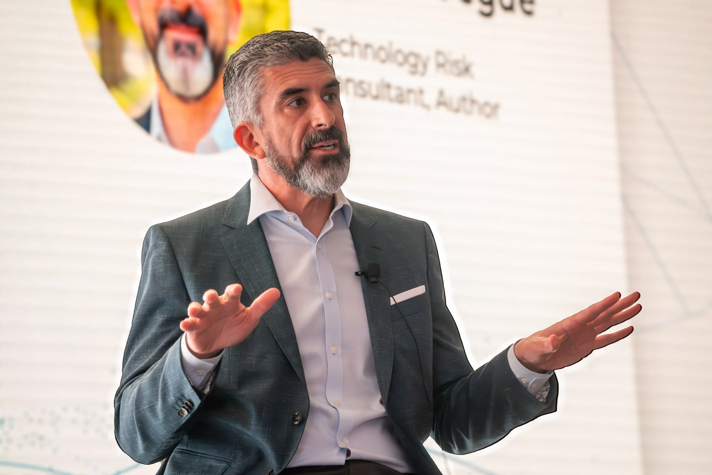 A man with gray hair and beard wearing a dark suit and white shirt, speaking and gesturing during a presentation in front of a screen with partially visible text.