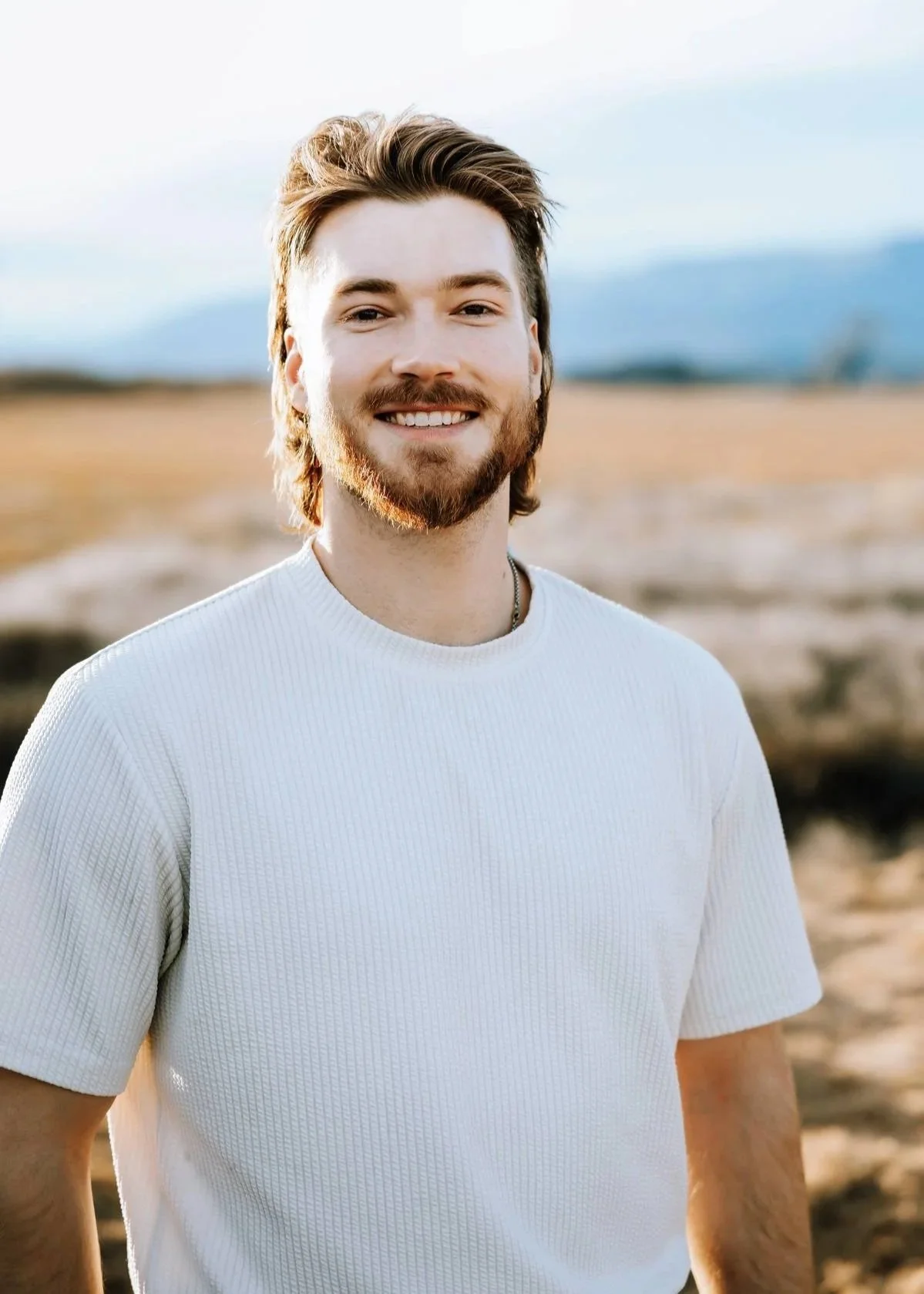 Man in a dark patterned shirt standing on a beach with the ocean and a distant rock formation in the background.