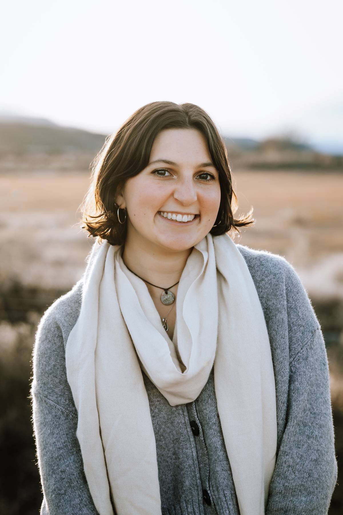 A woman in a light dress holding sunflowers, standing against a wooden wall, smiling.