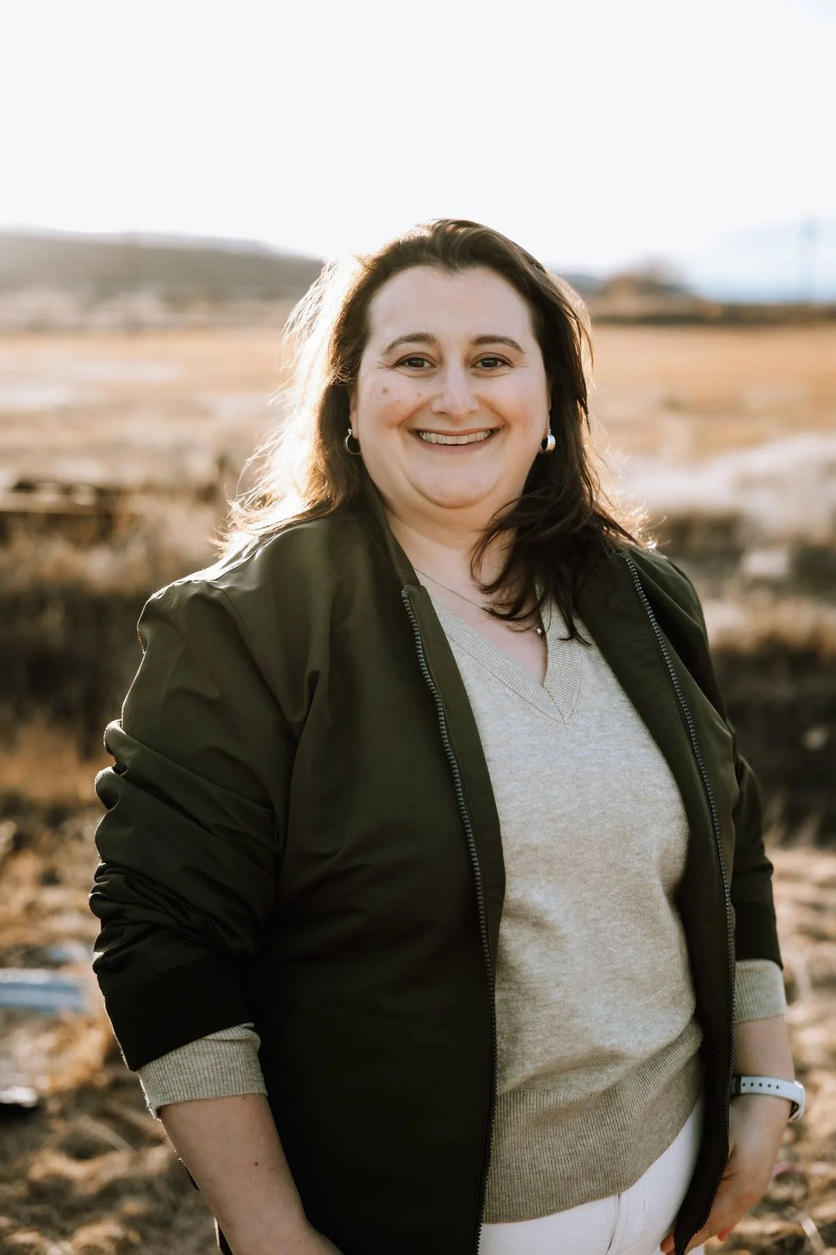 Person in a white blazer with a blue shirt, standing outside in front of a building and greenery.
