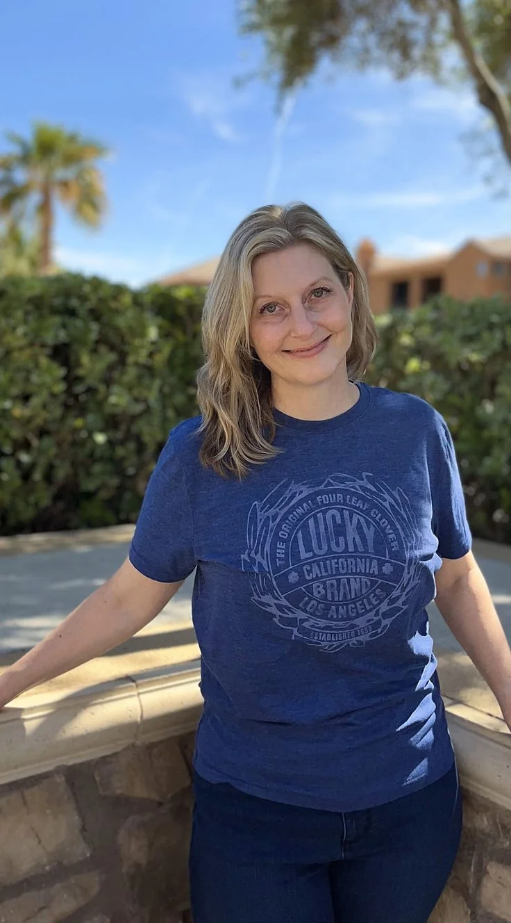 Woman with blonde wavy hair in a blue shirt stands outside smiling with green shrubs in the background.