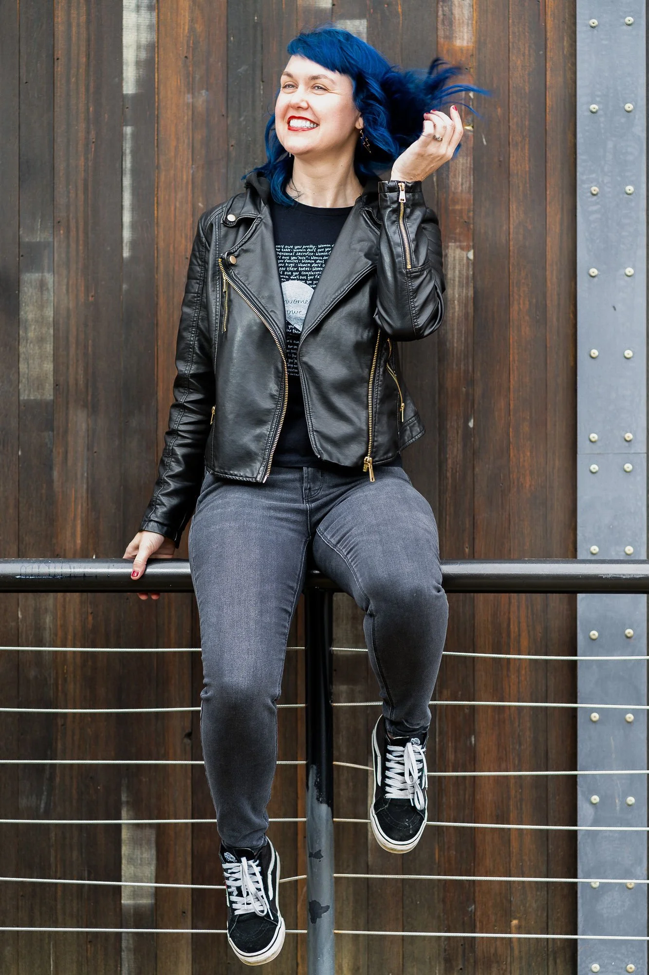 Woman with blue hair, a leather jacket, grey jeans and black sneakers sits on a metal railing, smiling and tossing her hair.