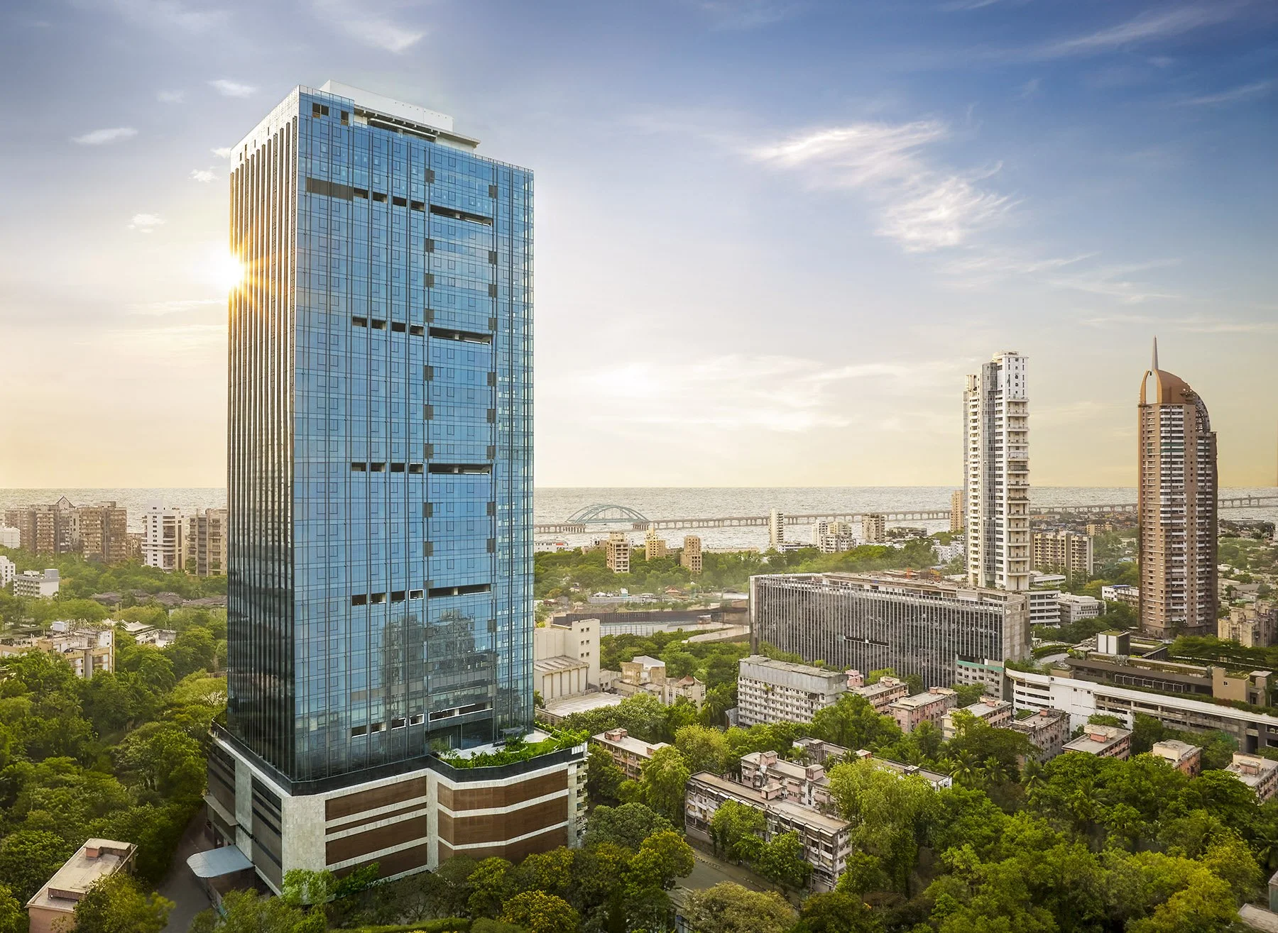 A cityscape featuring tall modern buildings, including a prominent blue glass skyscraper, with green trees in the foreground and a body of water in the background during sunset.