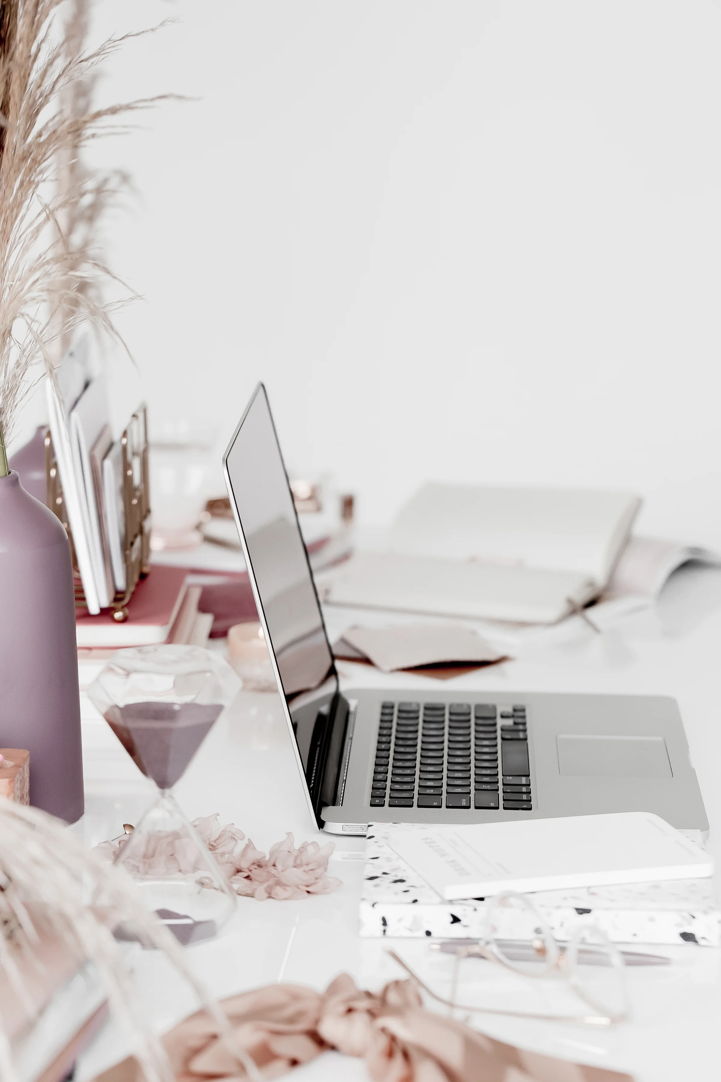 A desk with a silver laptop, some books, pink decorative items, and a tall vase with dried pampas grass on the left. There is a glass of wine and various paper decorations in the foreground.