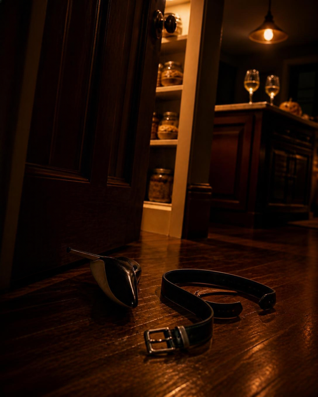 A woman's black high heel shoe and a man's black leather belt are on a hardwood floor in a dimly lit room, with a wooden cabinet, glass jars, wine glasses, and a ceiling light in the background.