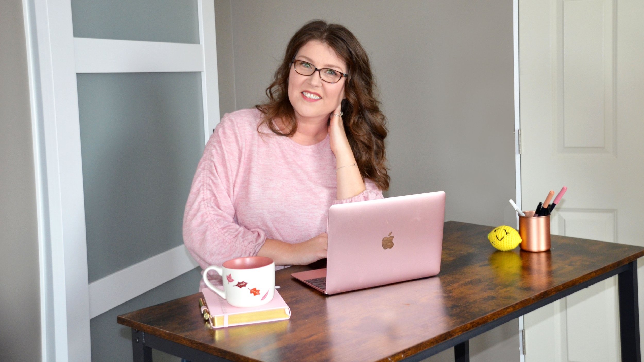 A woman with glasses and curly brown hair sitting at a desk with a pink laptop, smiling, wearing a pink long-sleeve shirt. The desk has a pink mug, a yellow notebook, a cup with pens, a crocheted yellow lemon, and a white door in the background.